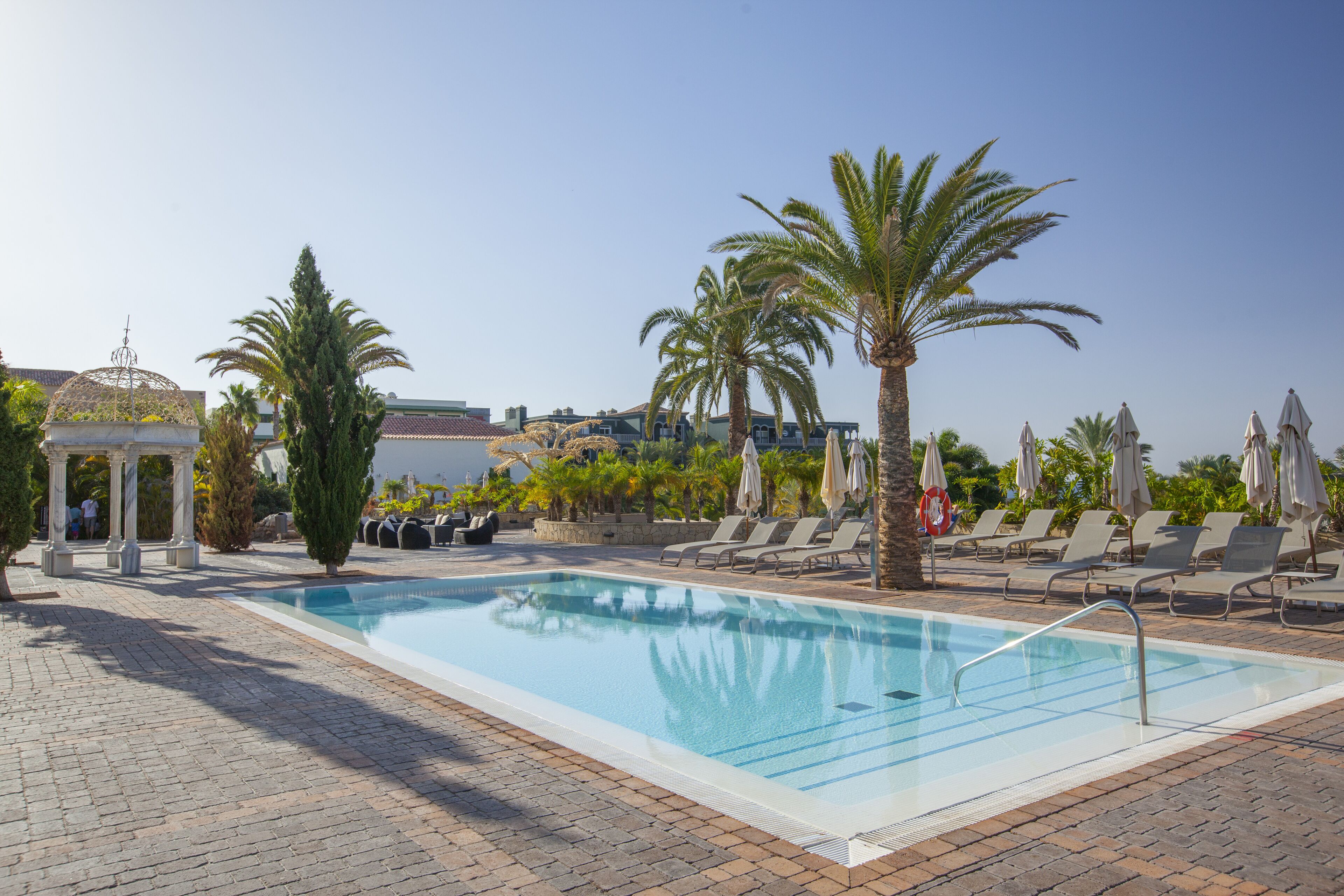 A pool area with lounge chairs, palm trees, and a gazebo.