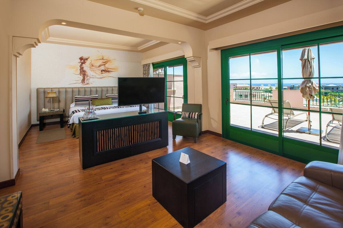 A spacious hotel room with a large bed, a TV, a small table, and a balcony with a view.