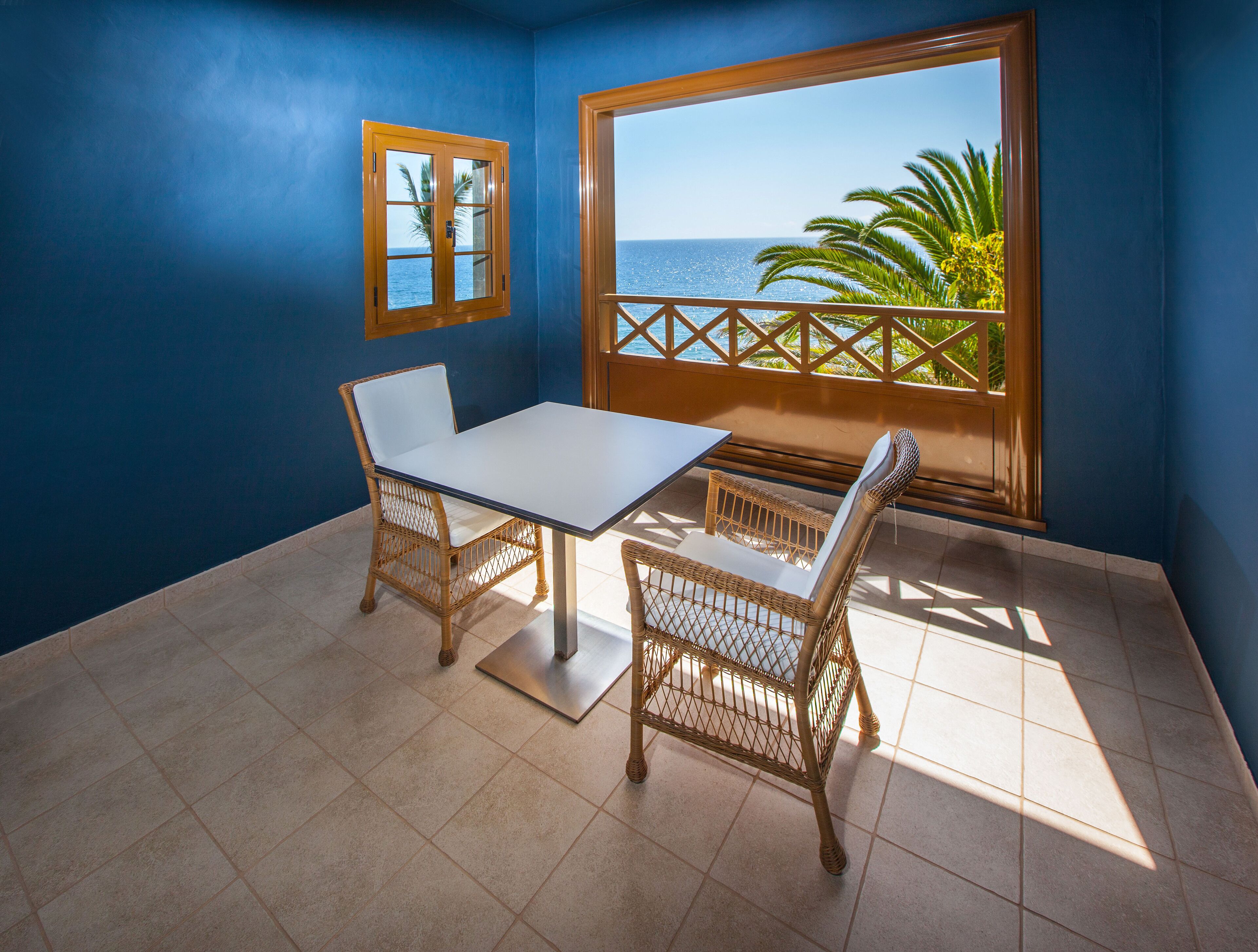 A balcony with a white table and wicker chairs, a view of the ocean, and a blue wall.