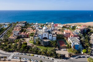 Vista aérea de un pueblo costero con una iglesia blanca destacada con cúpulas, rodeado de edificios residenciales y palmeras.