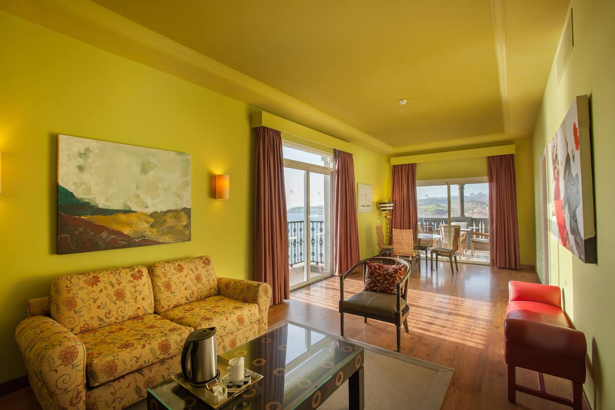 A living room with a sofa, armchair, and a coffee table. There is a balcony with a view.