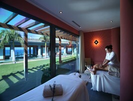 A spa room with a massage table, a towel, and a large window overlooking a pool and outdoor area.