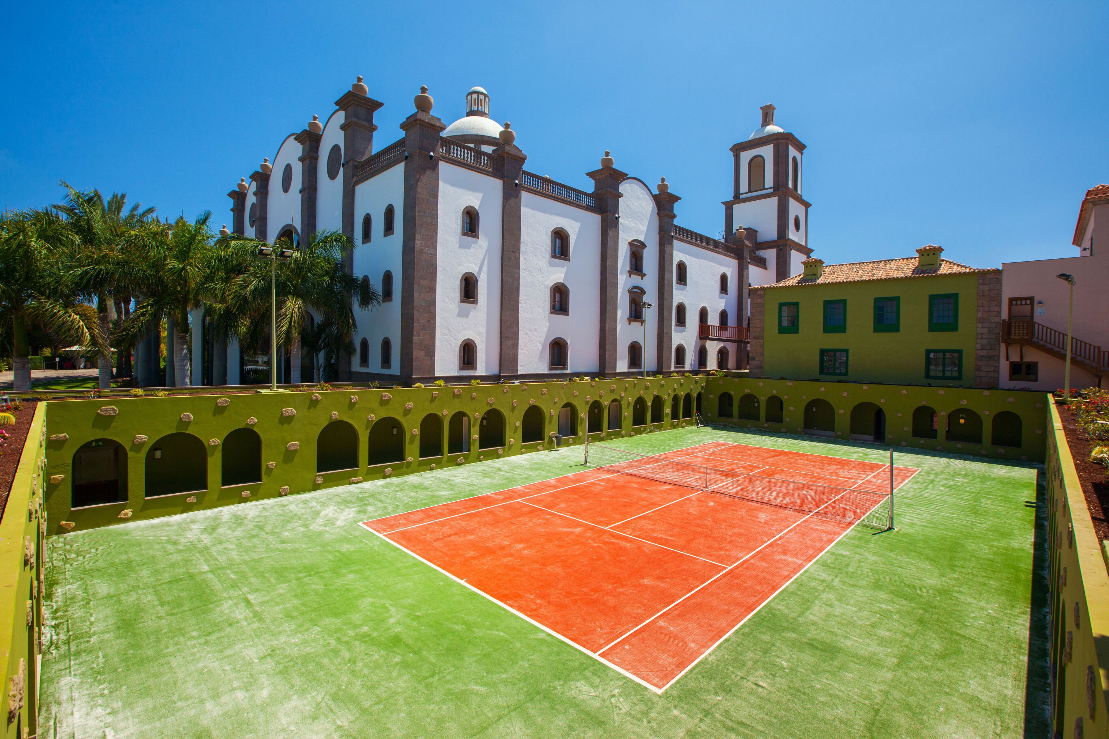 Ein Tennisplatz mit grün-orangefarbener Belag, umgeben von einer grünen Mauer mit Bögen, und ein weißes Gebäude mit einem Glockenturm im Hintergrund.