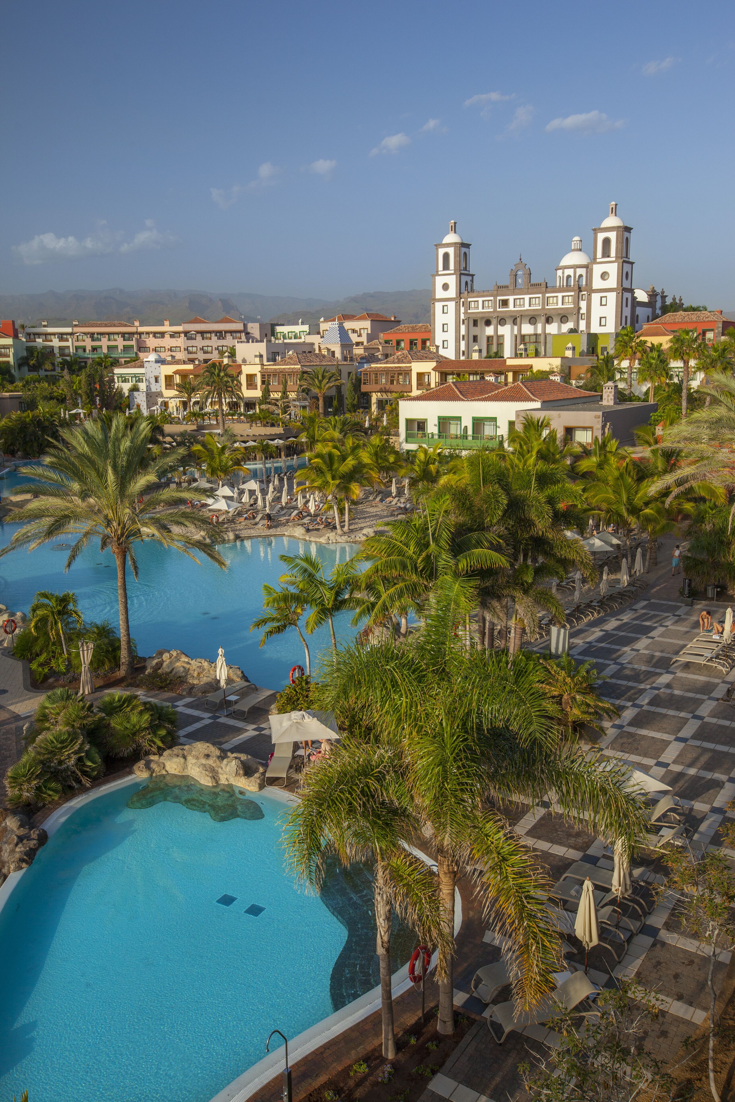 A resort with a large swimming pool surrounded by palm trees and lounge chairs, a church-like building in the background, and a mountainous landscape.