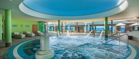 A large indoor pool with a central fountain, surrounded by lounge chairs and a view of the sea.