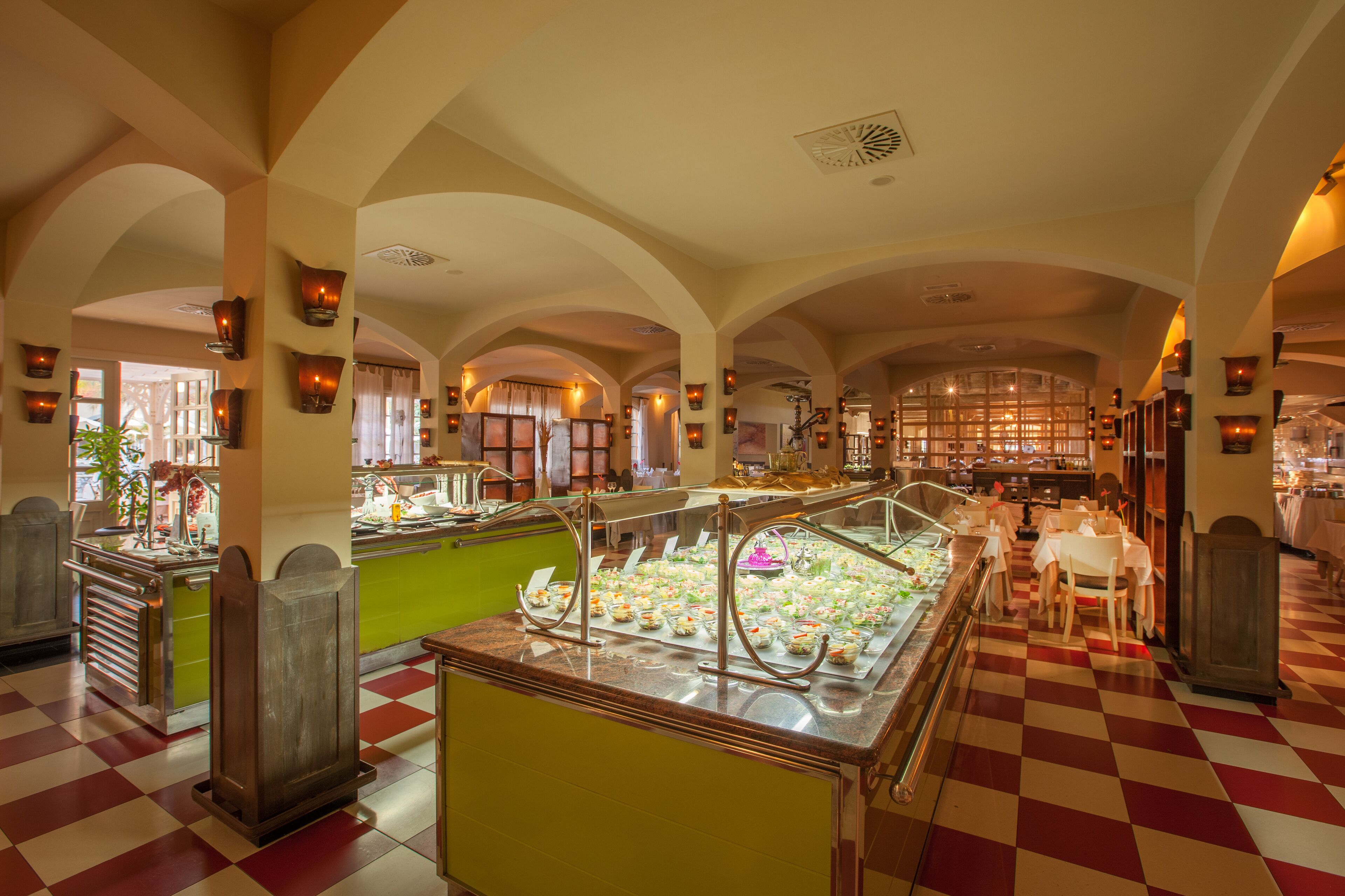 Un espace buffet doté d’un sol à carreaux, de plusieurs points de service où différents plats sont proposés, ainsi qu’un coin repas avec des tables et des chaises.