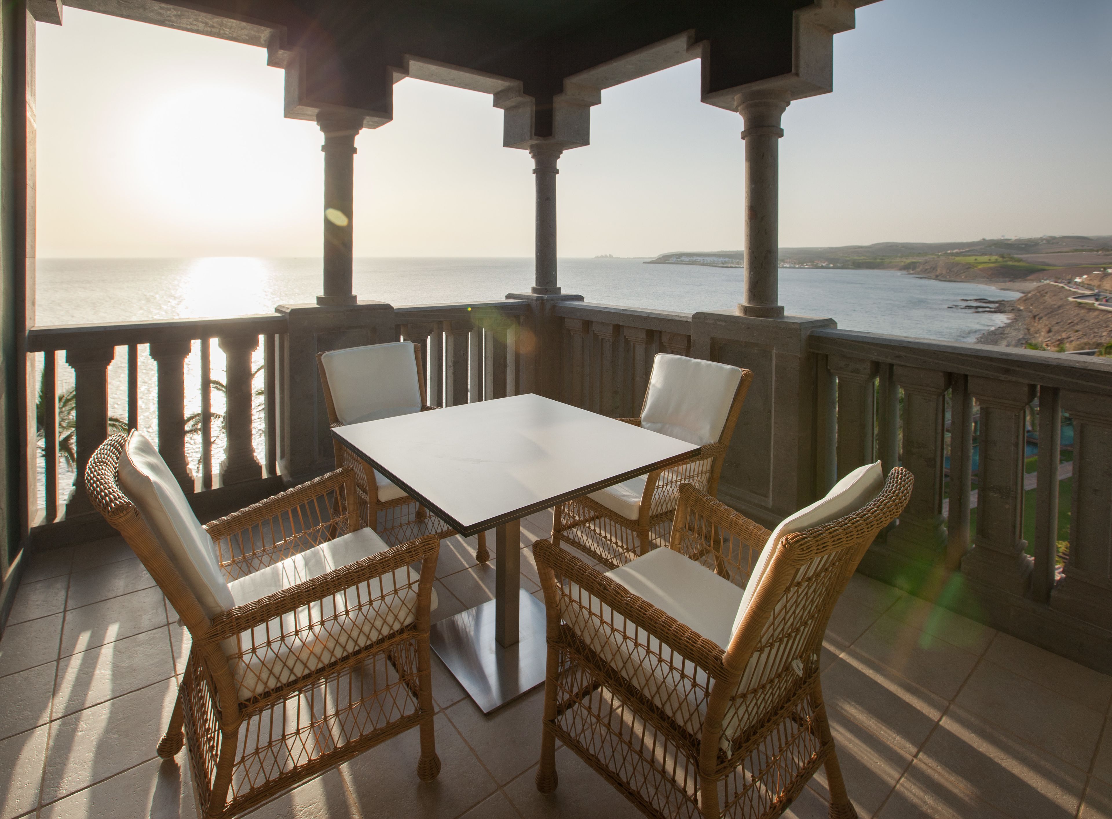 A covered outdoor seating area with a table and wicker chairs, overlooking a coastal landscape.
