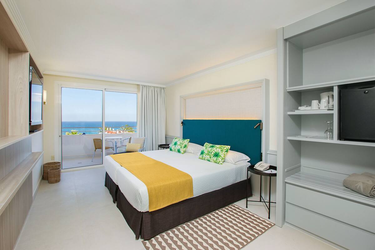 A hotel room with a bed, a desk, a chair, a TV, and a view of the sea.