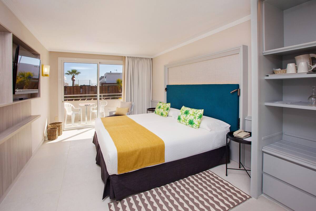 A hotel room with a large bed, a TV mounted on the wall, a balcony with outdoor seating, and a view of the cityscape.
