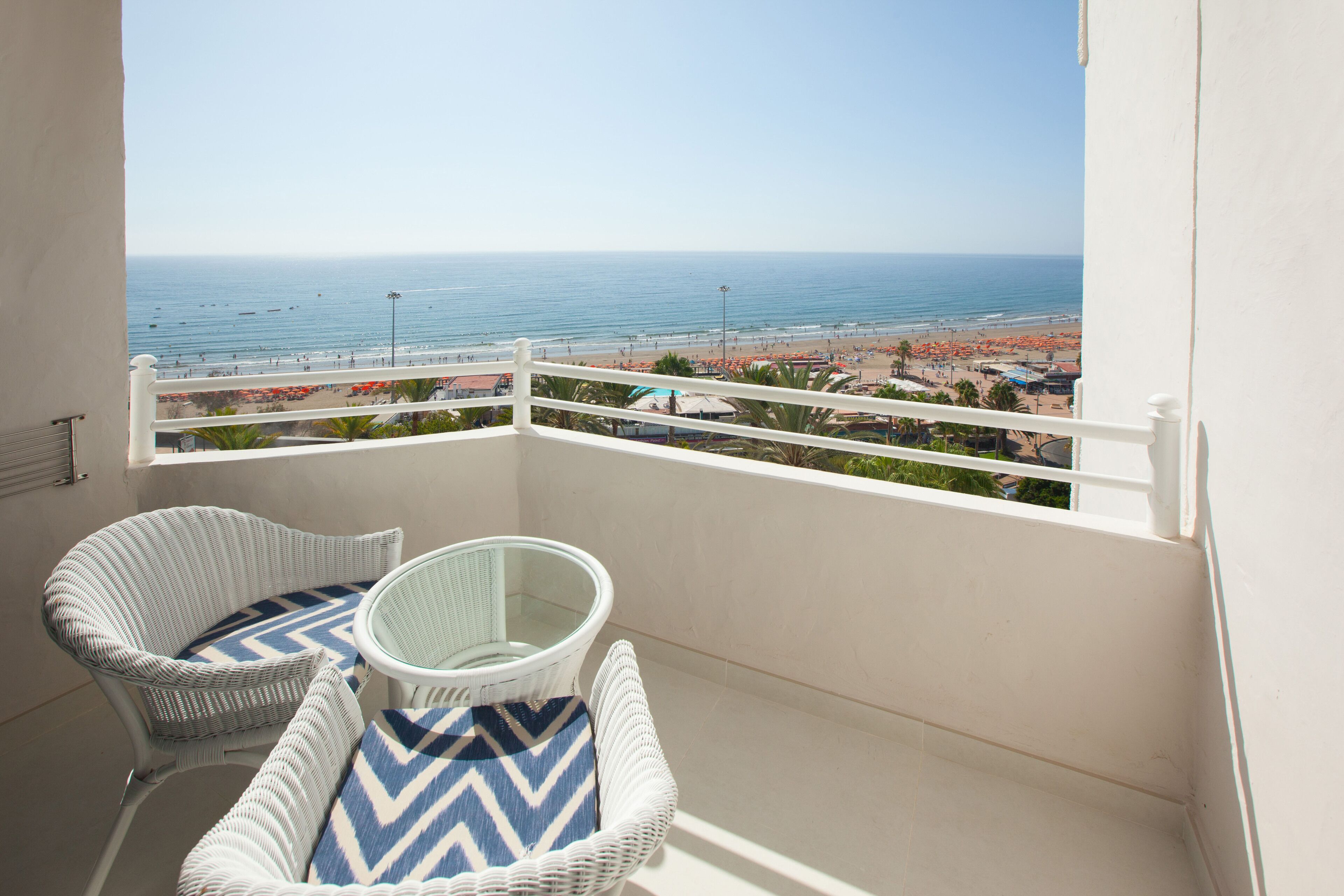 Ein Balkon mit Korbstühlen und einem Glastisch, mit Blick auf einen Strand mit Sonnenschirmen und Menschen.