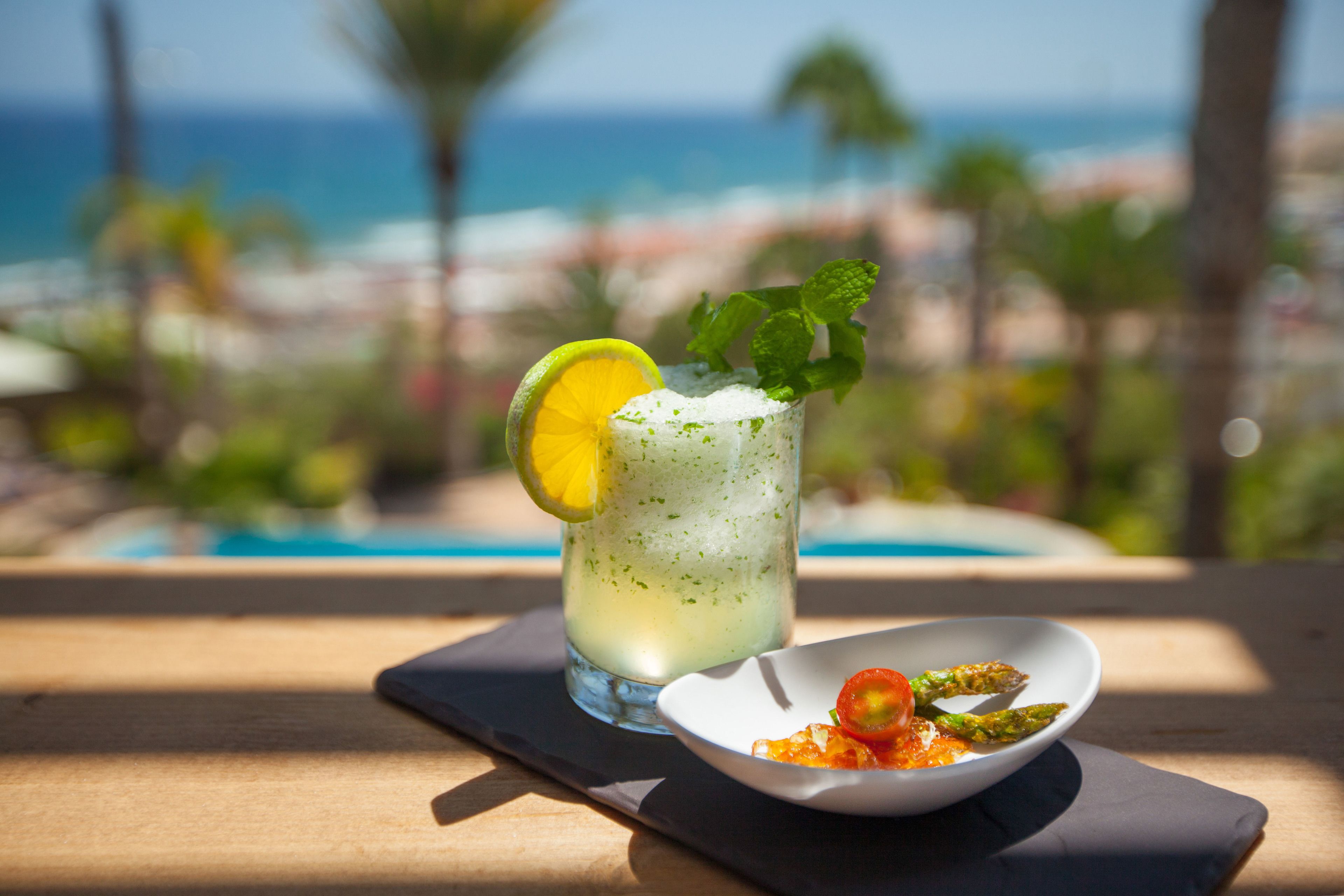 Ein Glas helles Getränk, garniert mit Limettenscheibe und Minzblättern, zusammen mit einer kleinen Portion Essen, vermutlich einem Salat, auf einem Holztisch. Im Hintergrund ist ein Pool und Palmen zu sehen.