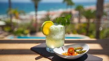 Ein Glas helles Getränk, garniert mit Limettenscheibe und Minzblättern, zusammen mit einer kleinen Portion Essen, vermutlich einem Salat, auf einem Holztisch. Im Hintergrund ist ein Pool und Palmen zu sehen.