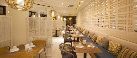 A modern restaurant with long communal tables, cushioned seating, and decorative lighting.