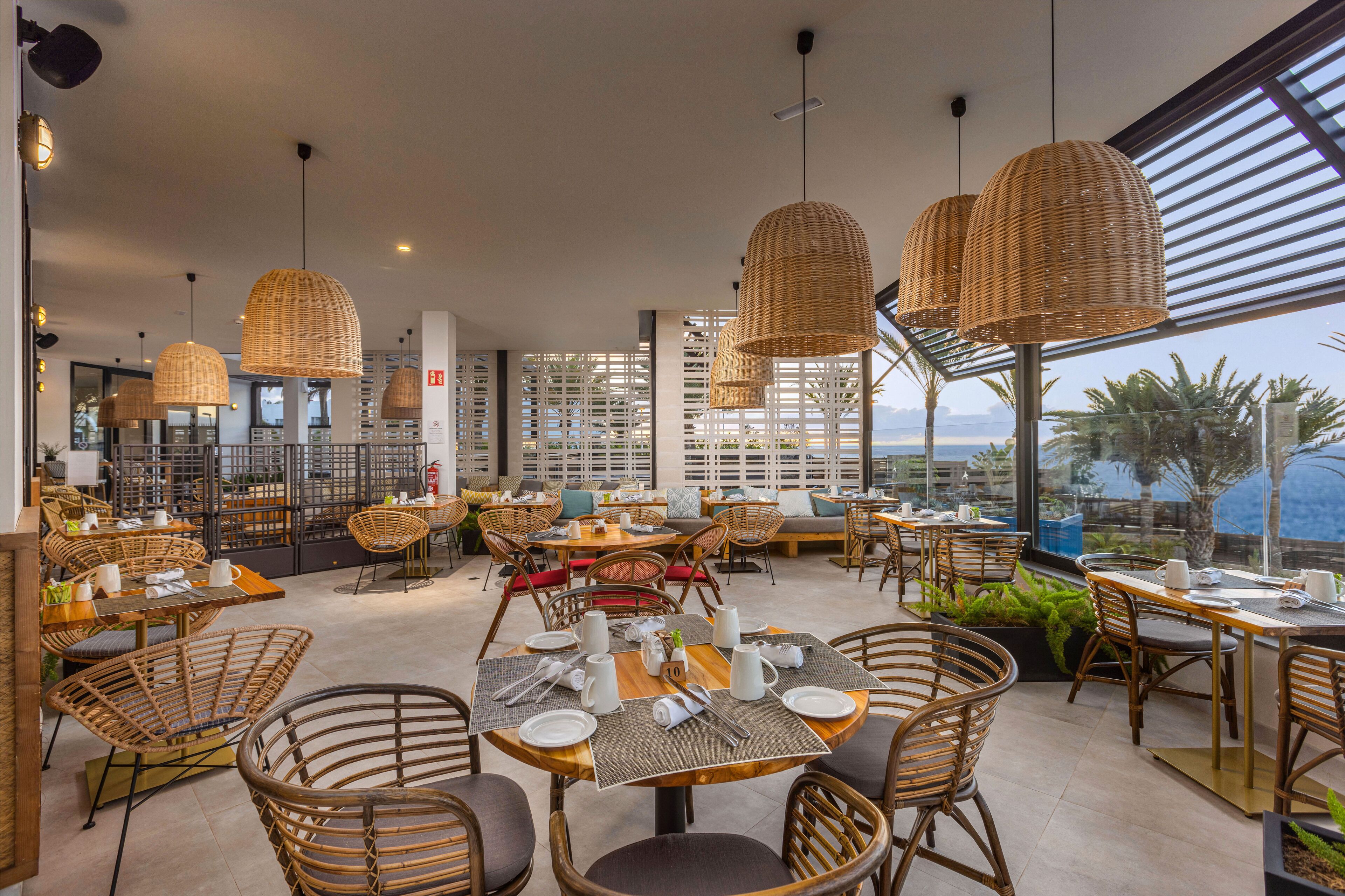 A restaurant with wicker furniture, round tables, and hanging wicker lamps.