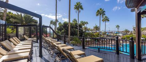 A deck with sun loungers overlooking a pool, surrounded by palm trees and a clear sky.