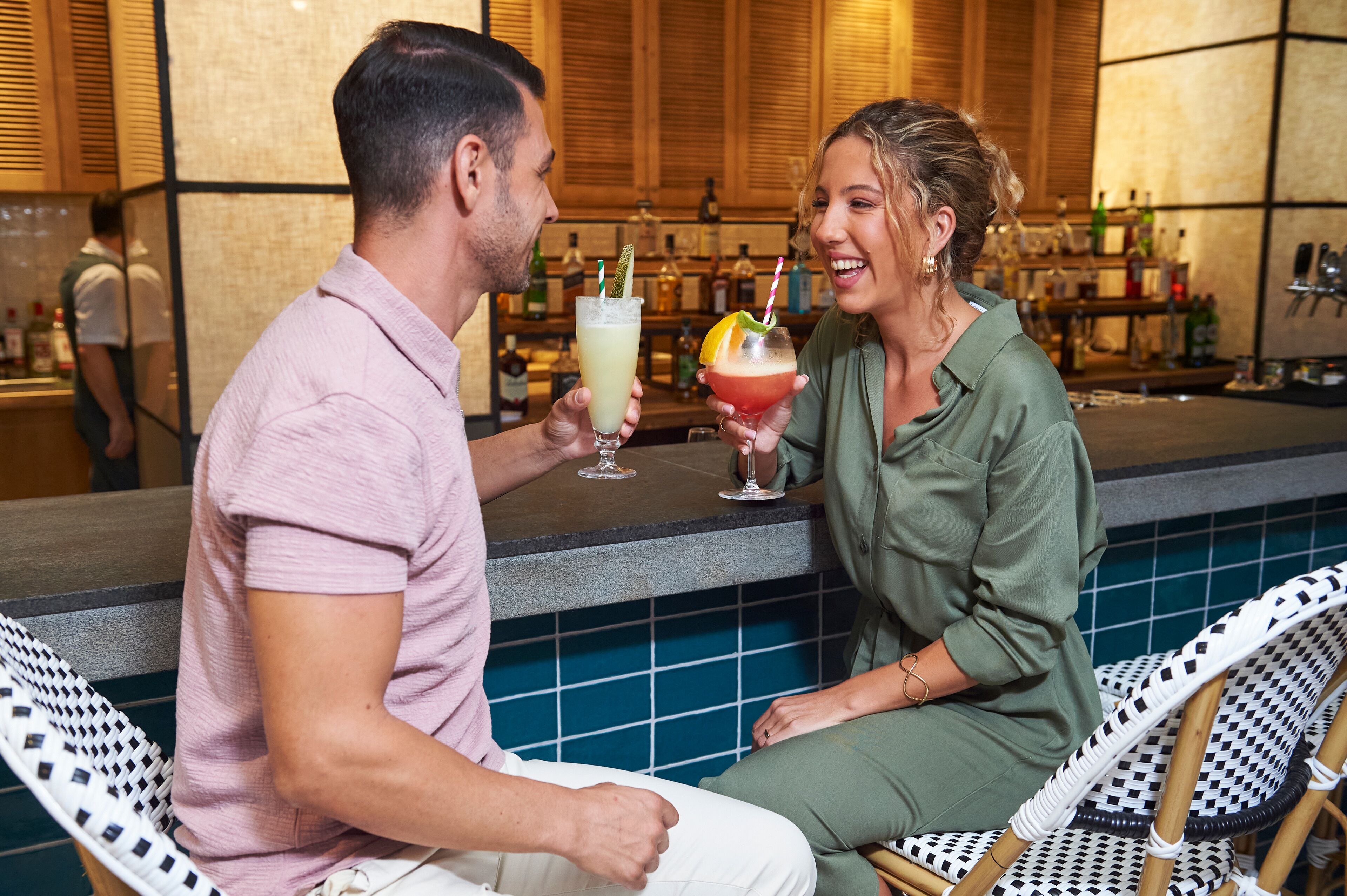 Una pareja sentada en la barra con bebidas, un reverso de azulejos y un cantinero al fondo.