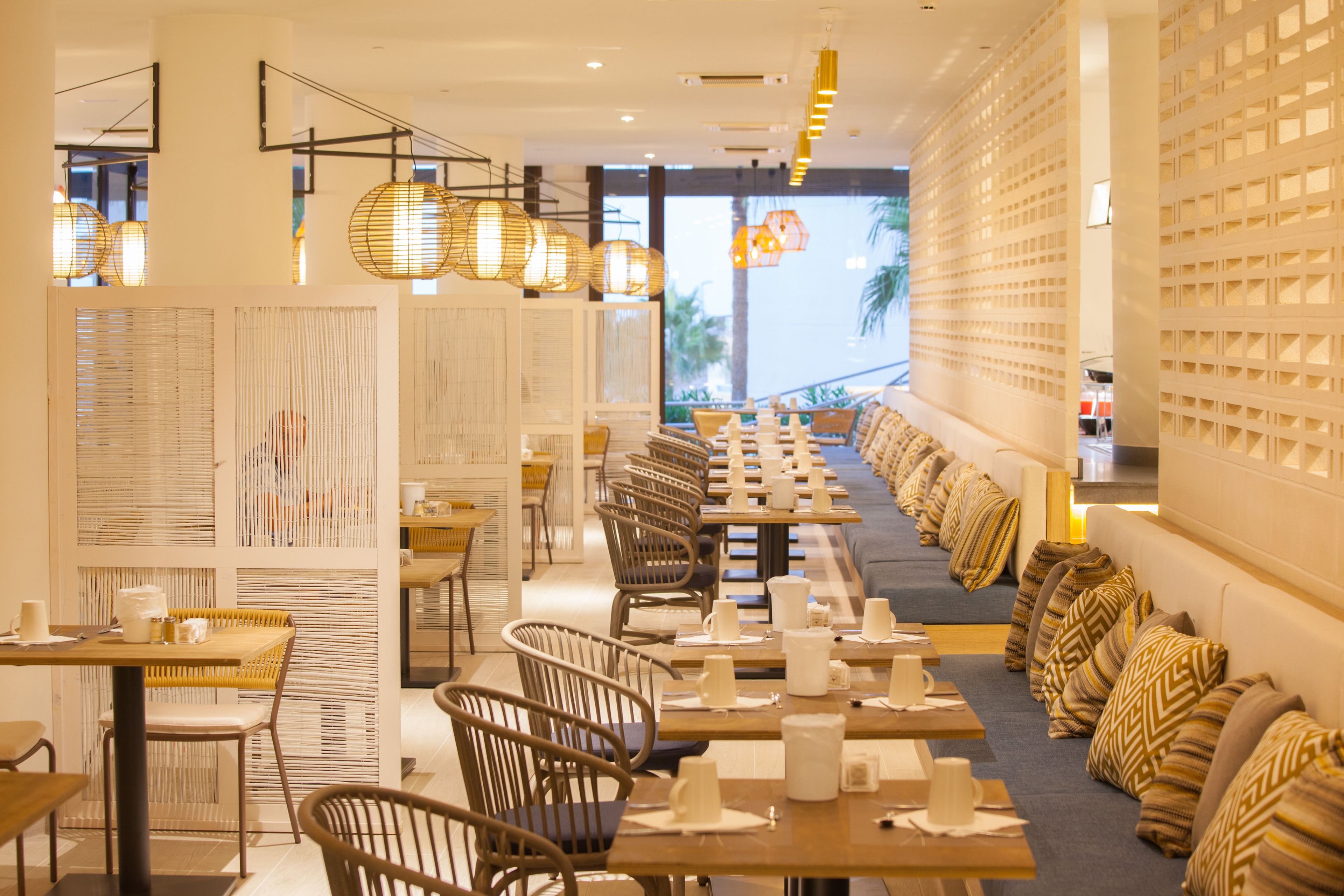 Ein Restaurant mit langen Gemeinschaftstischen, Korbstühlen und gemusterten Kissen.