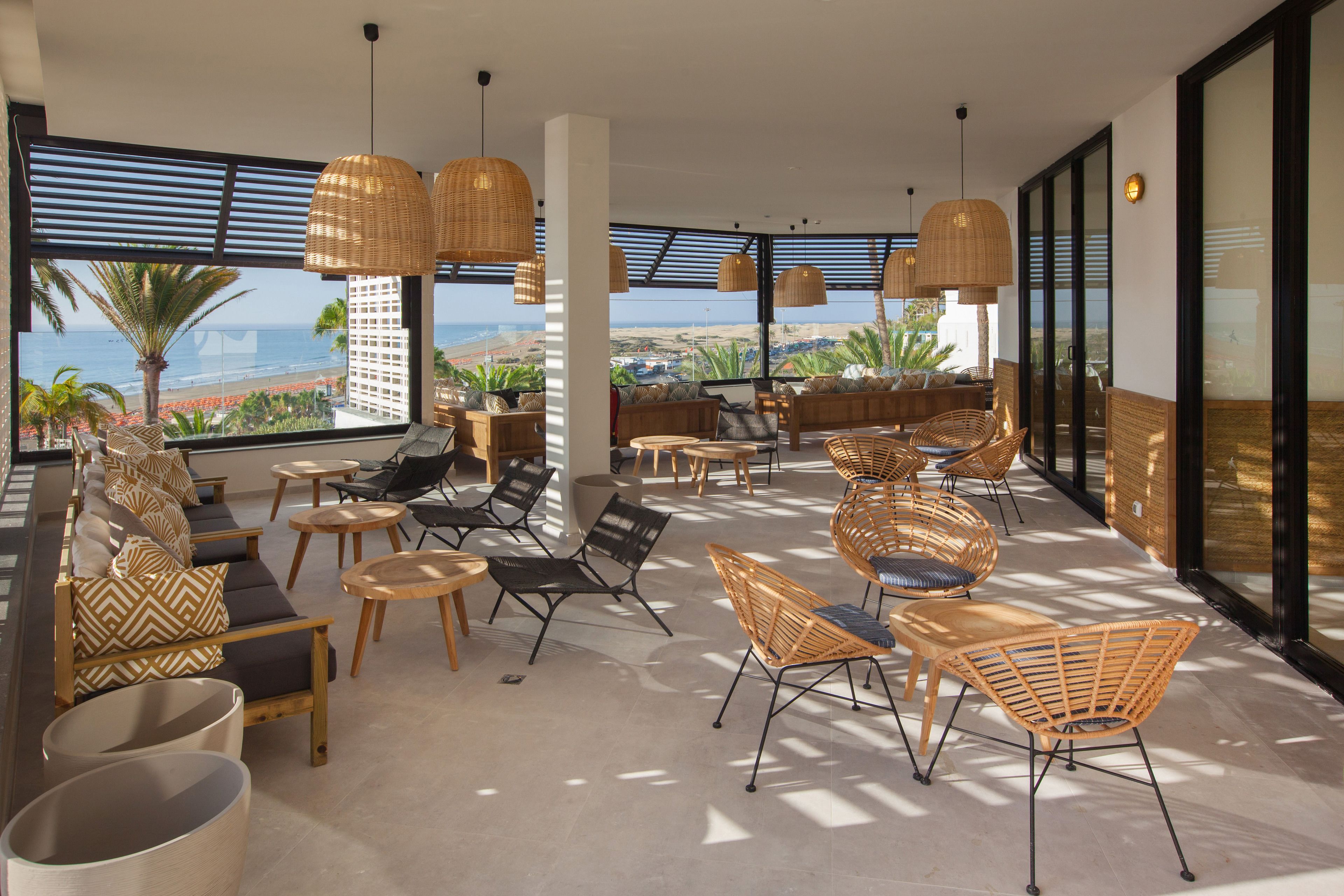 A spacious outdoor lounge area with wicker furniture, large windows offering a view of the beach and ocean, and a modern architectural design.