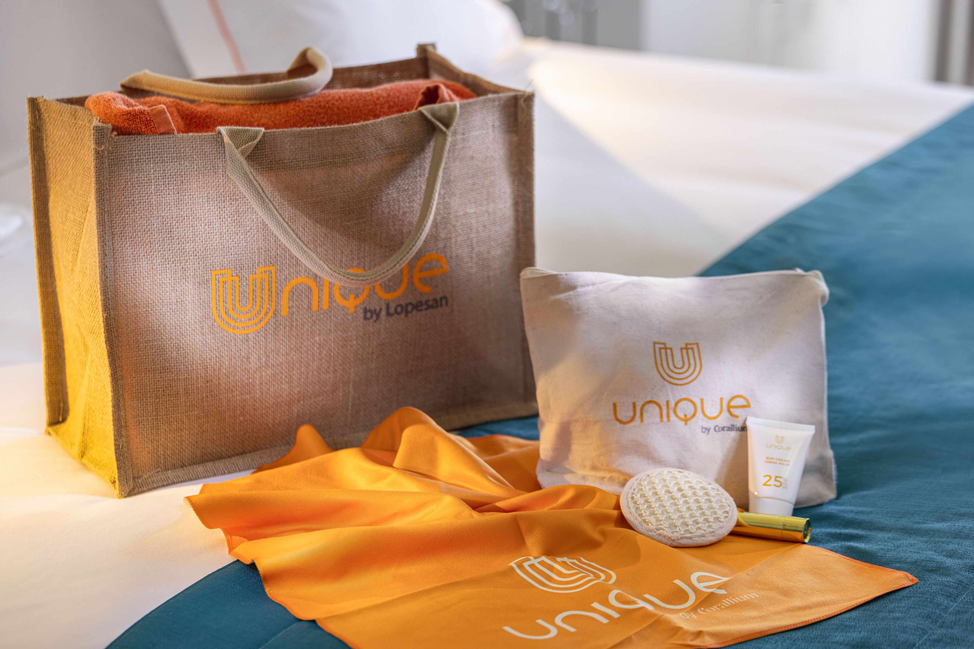 Une chambre d’hôtel avec un lit, une grande valise marron, une plus petite valise blanche, un tissu orange et une bouteille blanche avec une étiquette.