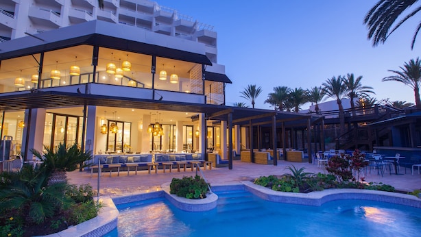 A modern hotel with a pool area, outdoor seating, and a well-lit lobby. - Corallium Dunamar by Lopesan Hotels - Adults Only (San Bartolomé de Tirajana)