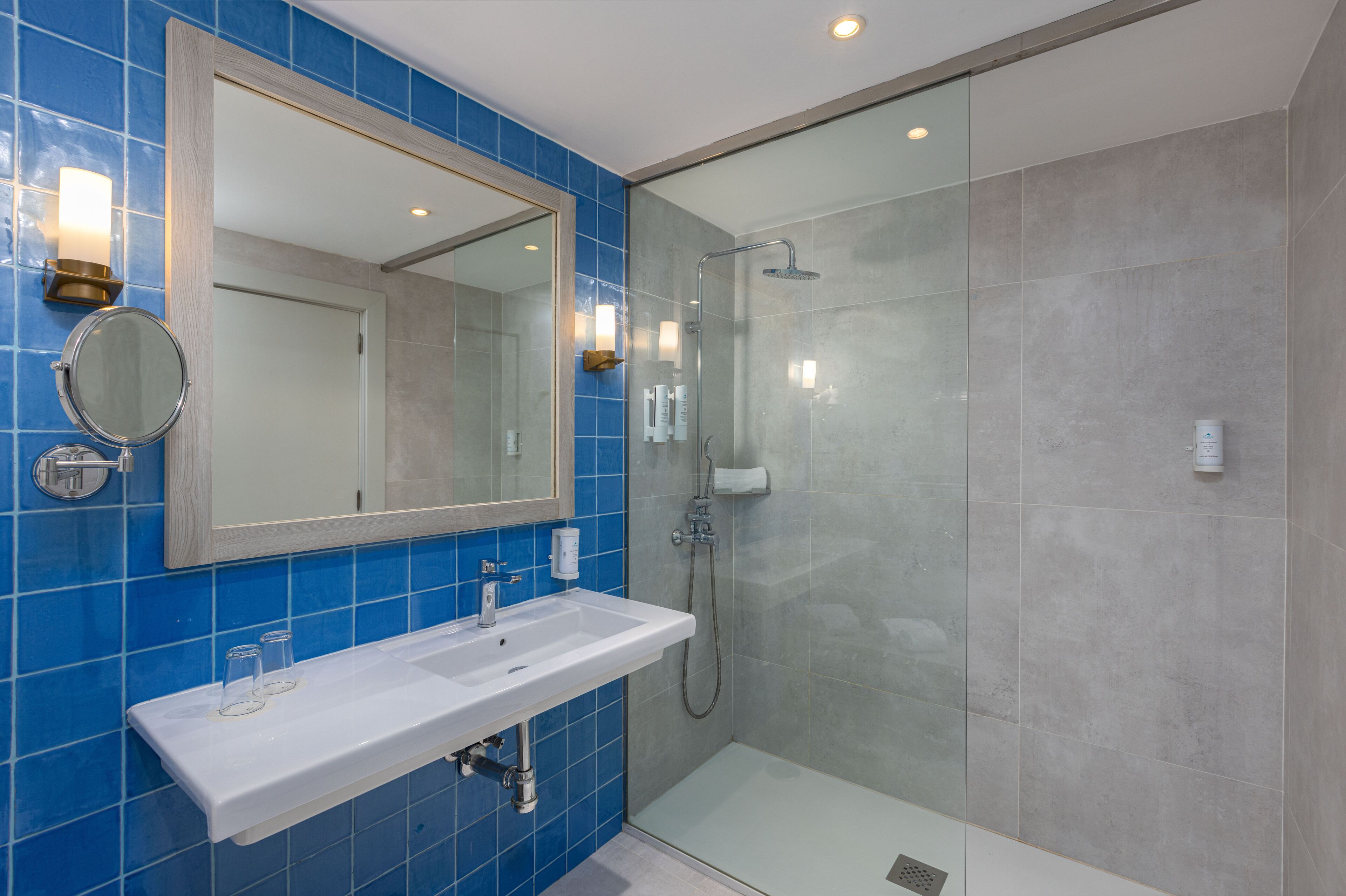 Een badkamer met een wit wastafelmeubel, blauwe tegels, een grote spiegel en een douche met glazen wanden.