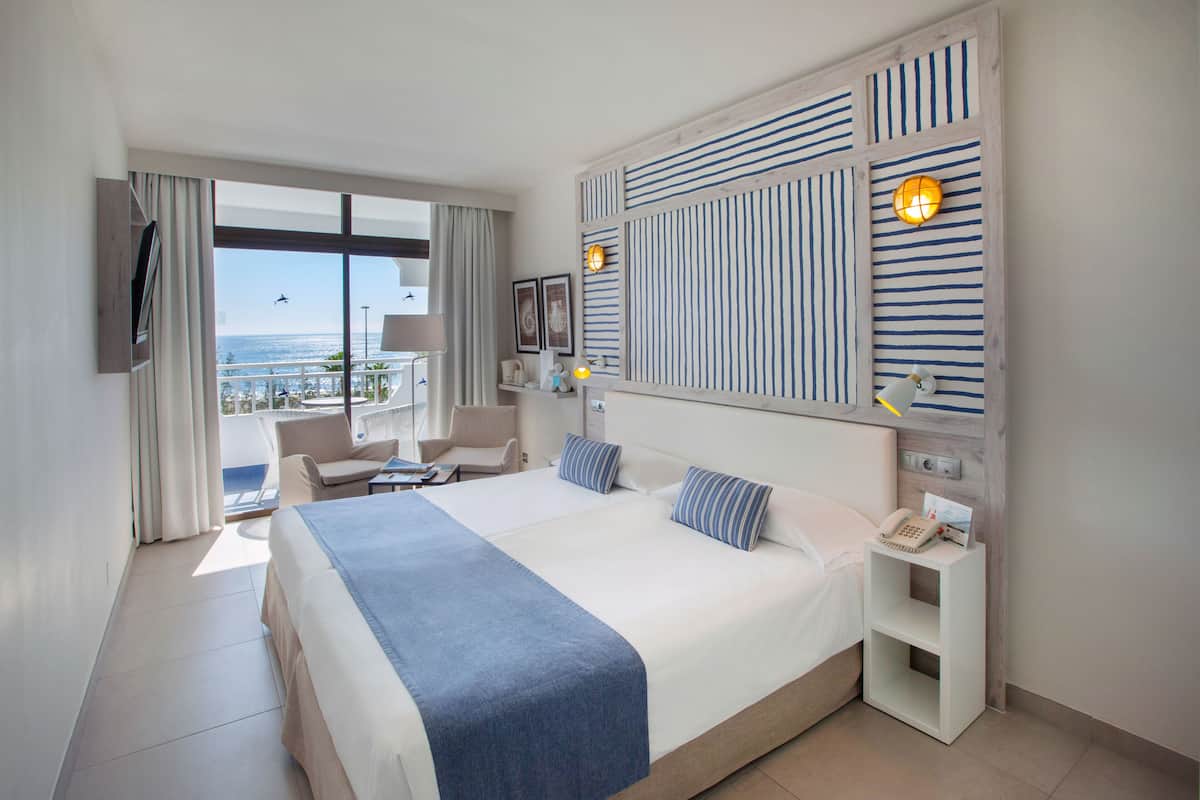 A hotel room with a large bed, two armchairs, a small table, a TV, a nightstand, and a view of the ocean.