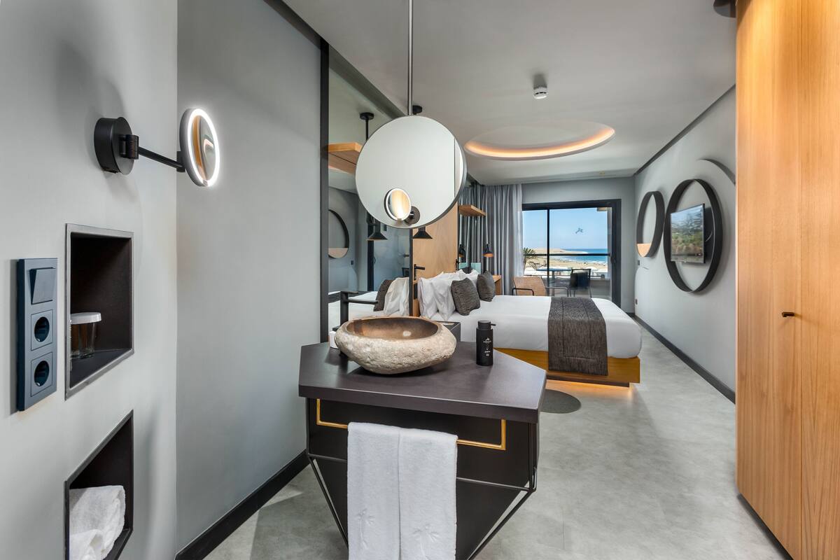 A modern hotel room with a large bed, a vanity with a bowl sink, a mirror, and a view of the beach through the window.