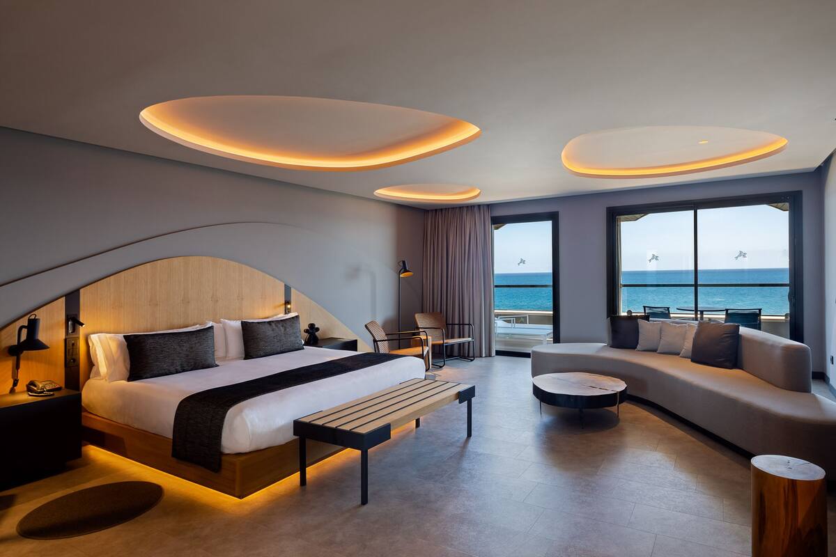 A modern hotel room with a curved bed, a sofa, a small table, and a view of the sea.