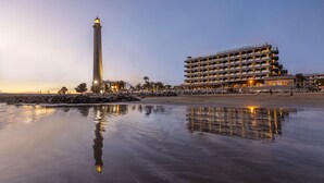 Un hotel frente al mar con un faro destacado, un edificio de varios pisos con balcones, y una superficie acuática tranquila y reflectante.