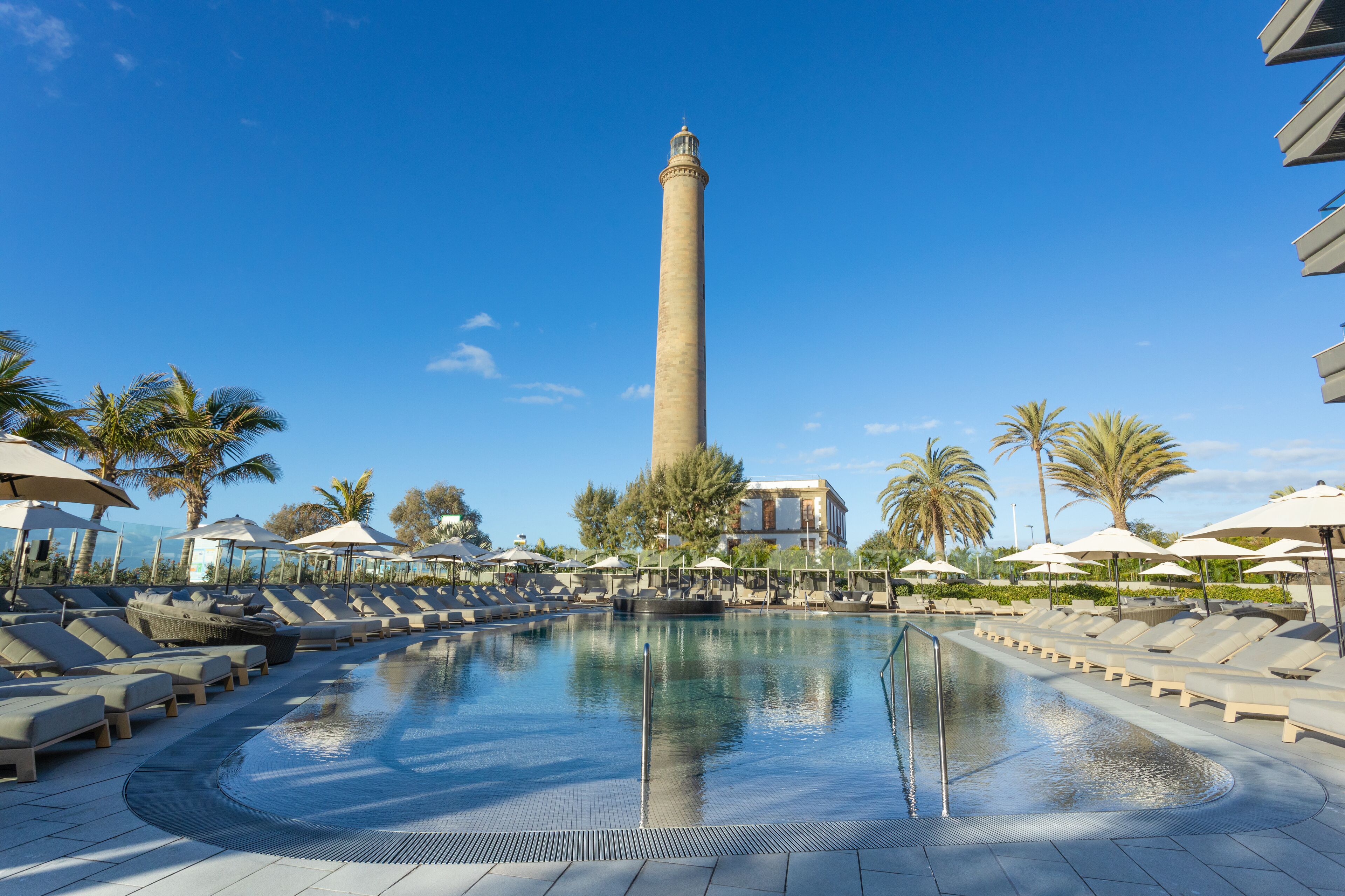 Zona de piscina con sillas de descanso y sombrillas, una torre alta al fondo y palmeras.