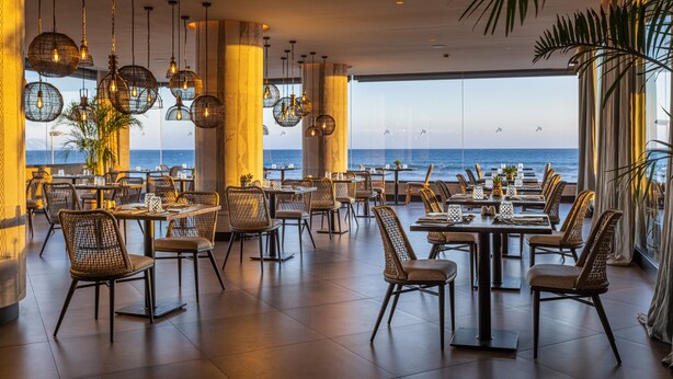 Un restaurante con mesas y sillas dispuestas para comer, amplias ventanas que dan a una vista del océano, y lámparas colgantes.