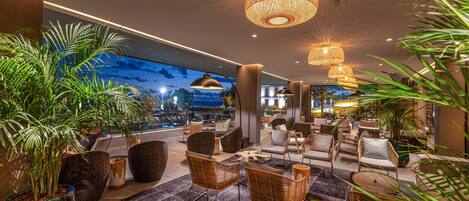 A modern lounge area with wicker furniture, potted plants, and a large window overlooking a cityscape.