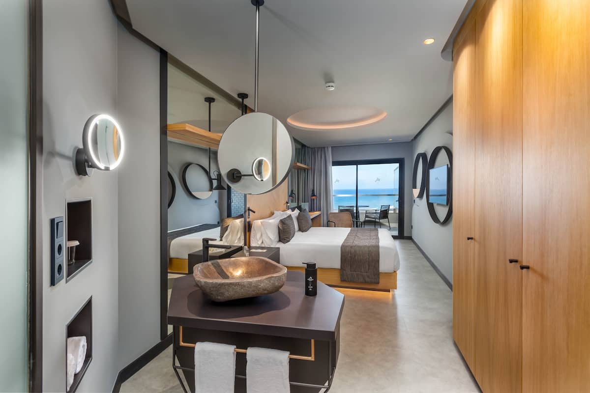 A modern hotel room with a large bed, a sitting area, a dining table with a bowl centerpiece, and a view of the sea.
