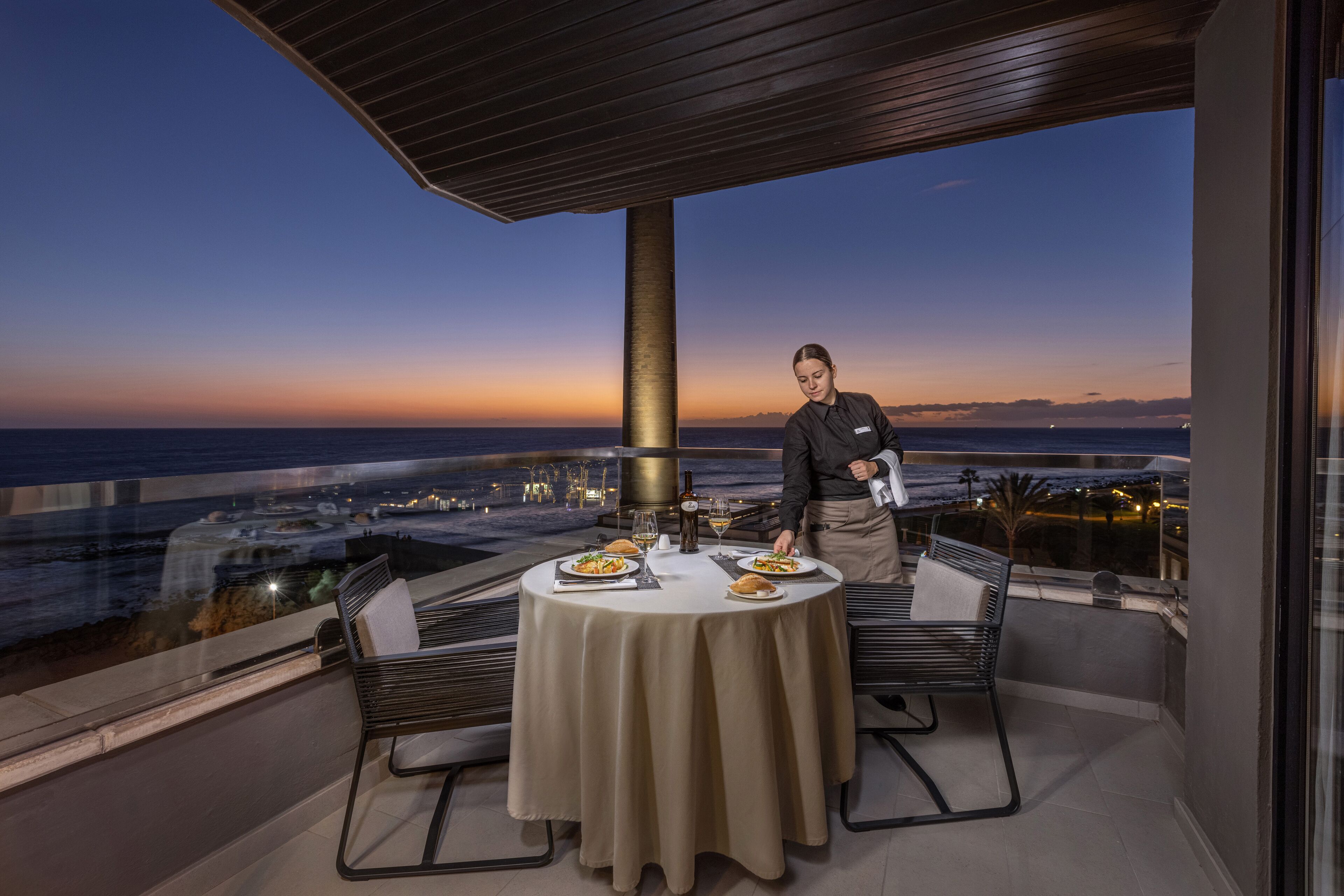 A dining area with a table set for two, overlooking a scenic ocean view at dusk.