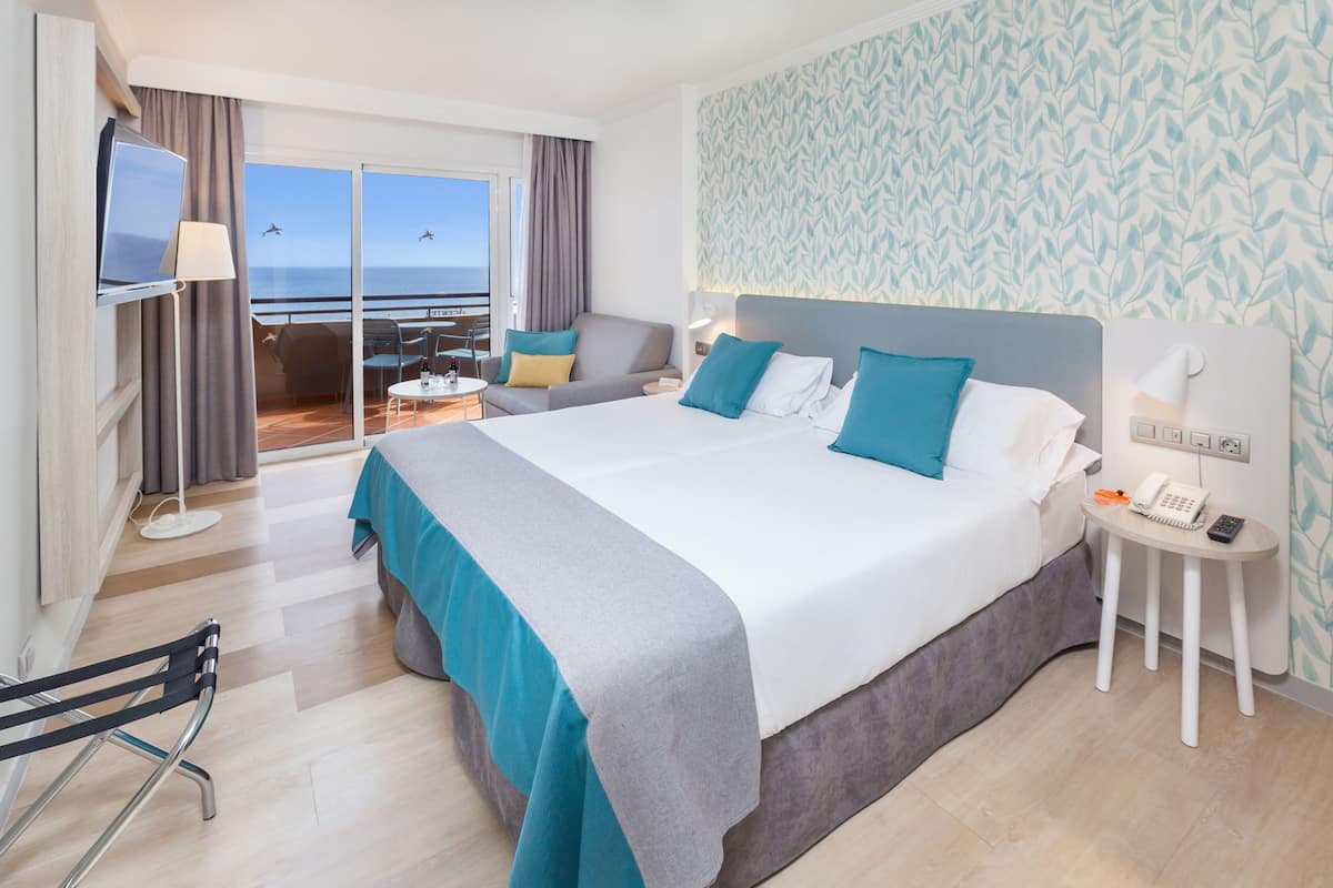 A hotel room with a large bed, a desk, a chair, a TV, and a view of the ocean.