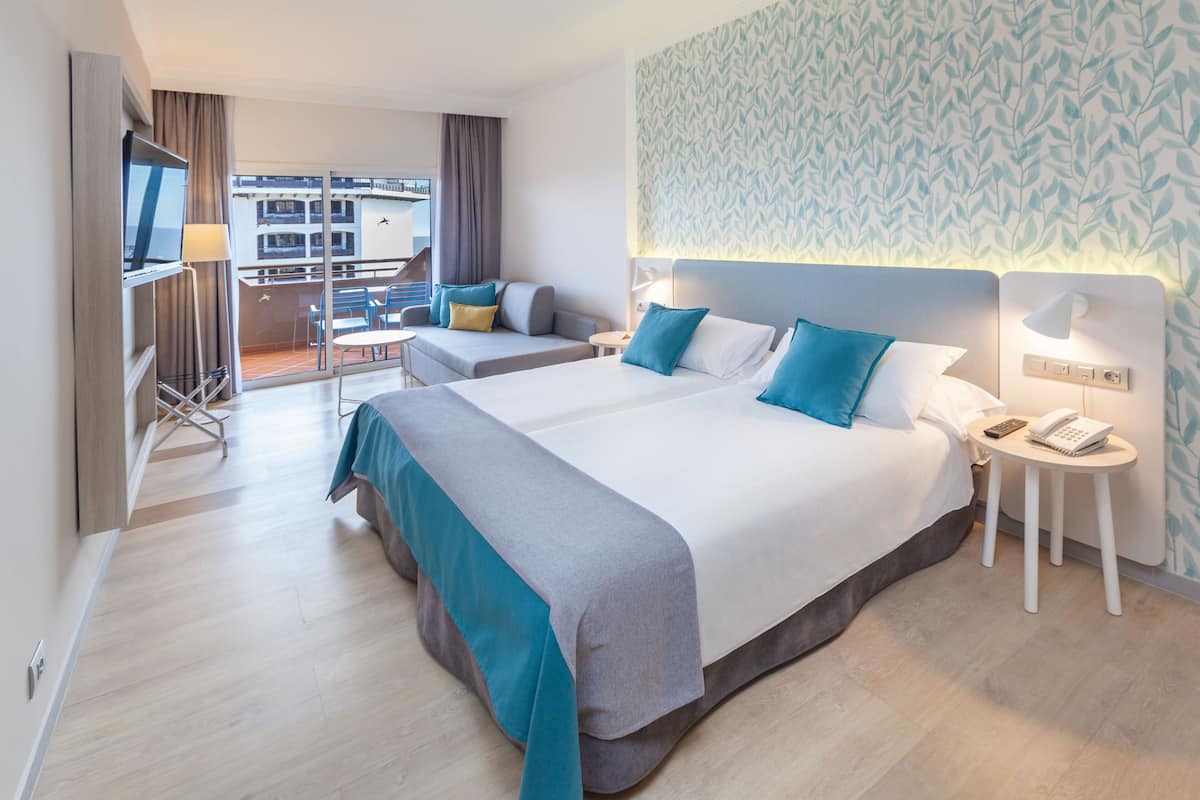 A hotel room with two beds, a TV, a desk, and a balcony with a view of buildings.