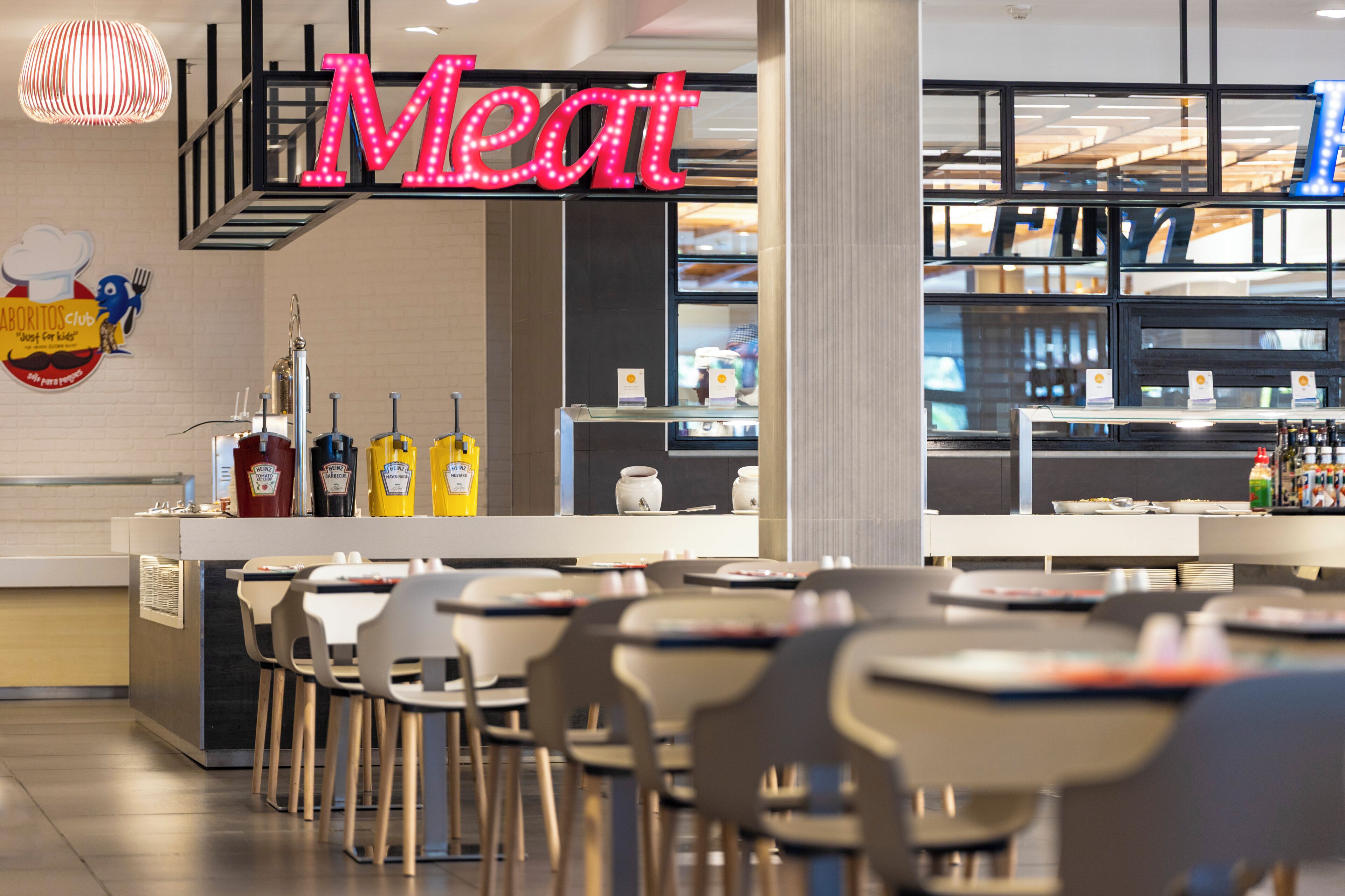 A restaurant interior with a neon sign reading 'Meat,' a counter with a display case, and several tables and chairs.