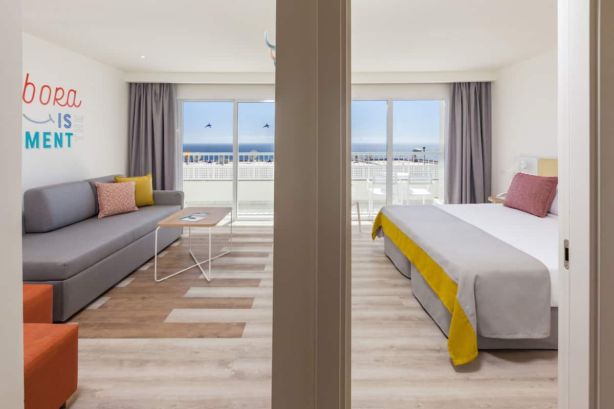 Une chambre d’hôtel moderne avec un canapé gris, un lit agrémenté d’une touche de jaune, une petite table et une vue sur la mer grâce à de grandes fenêtres.