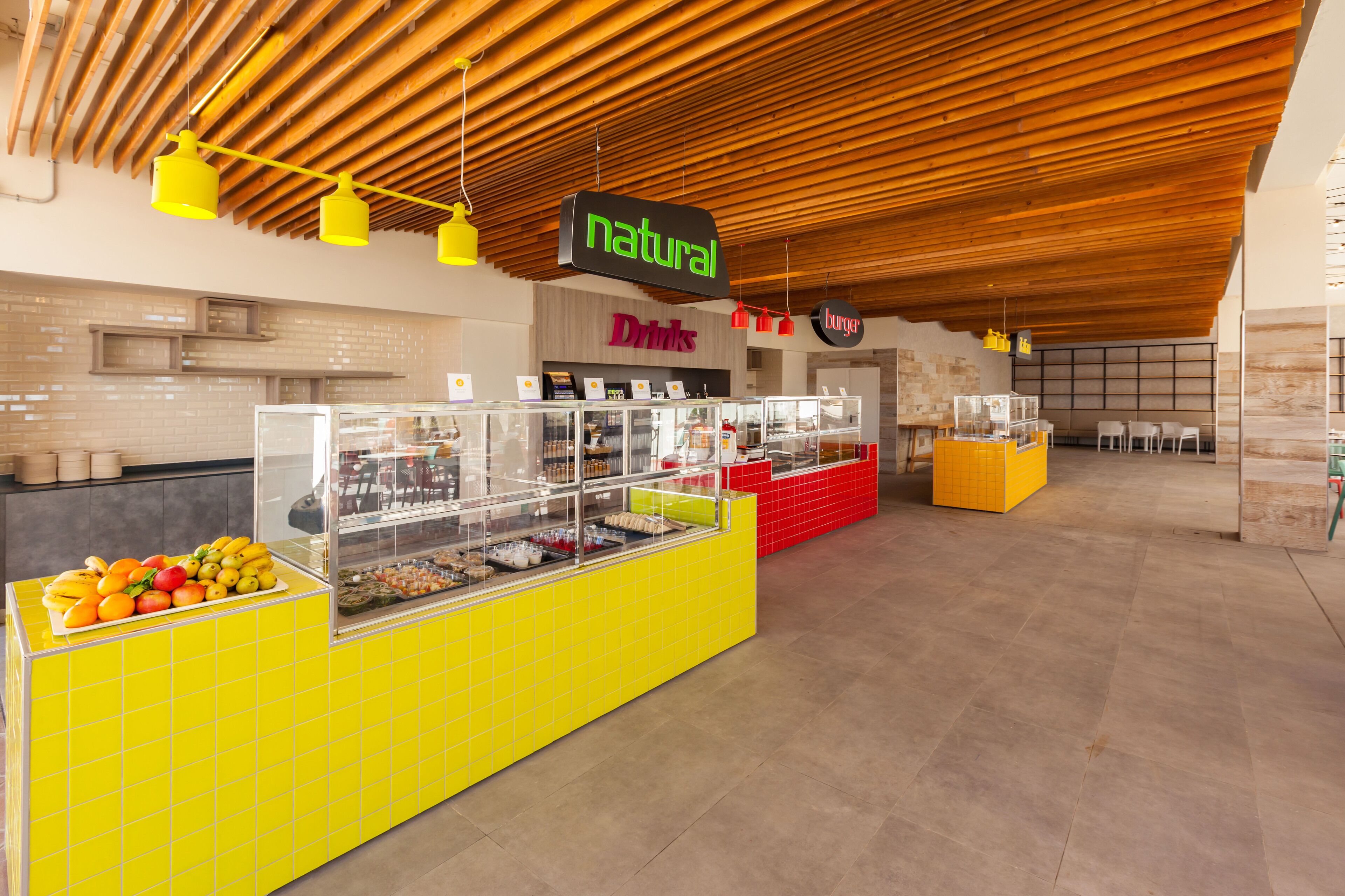 Un patio de comidas con un mostrador amarillo que exhibe frutas y un letrero que dice 'Natural Drinks'.