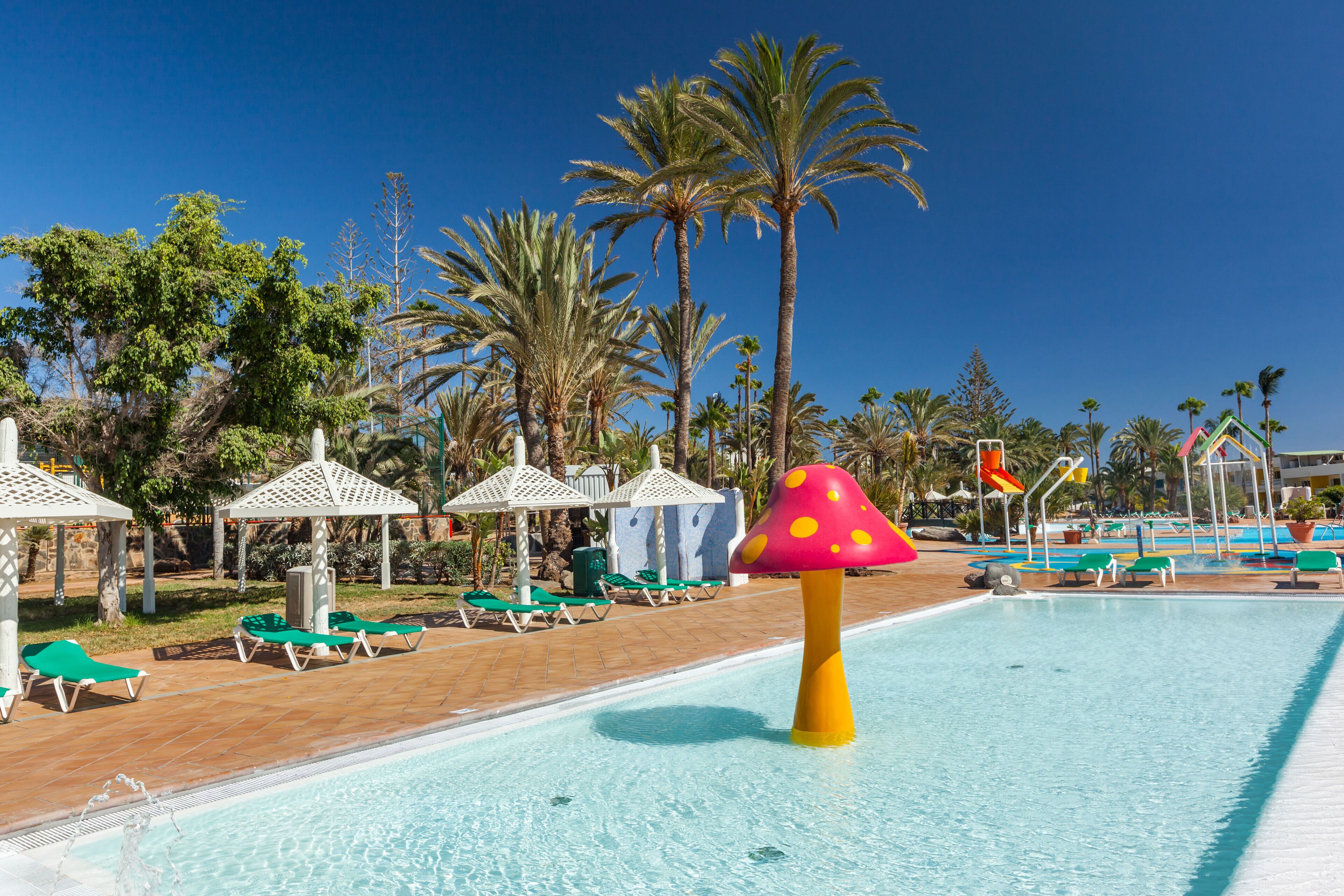 Een zwembadgedeelte met ligbedden, parasols en een opvallende waterattractie in de vorm van een paddenstoel.