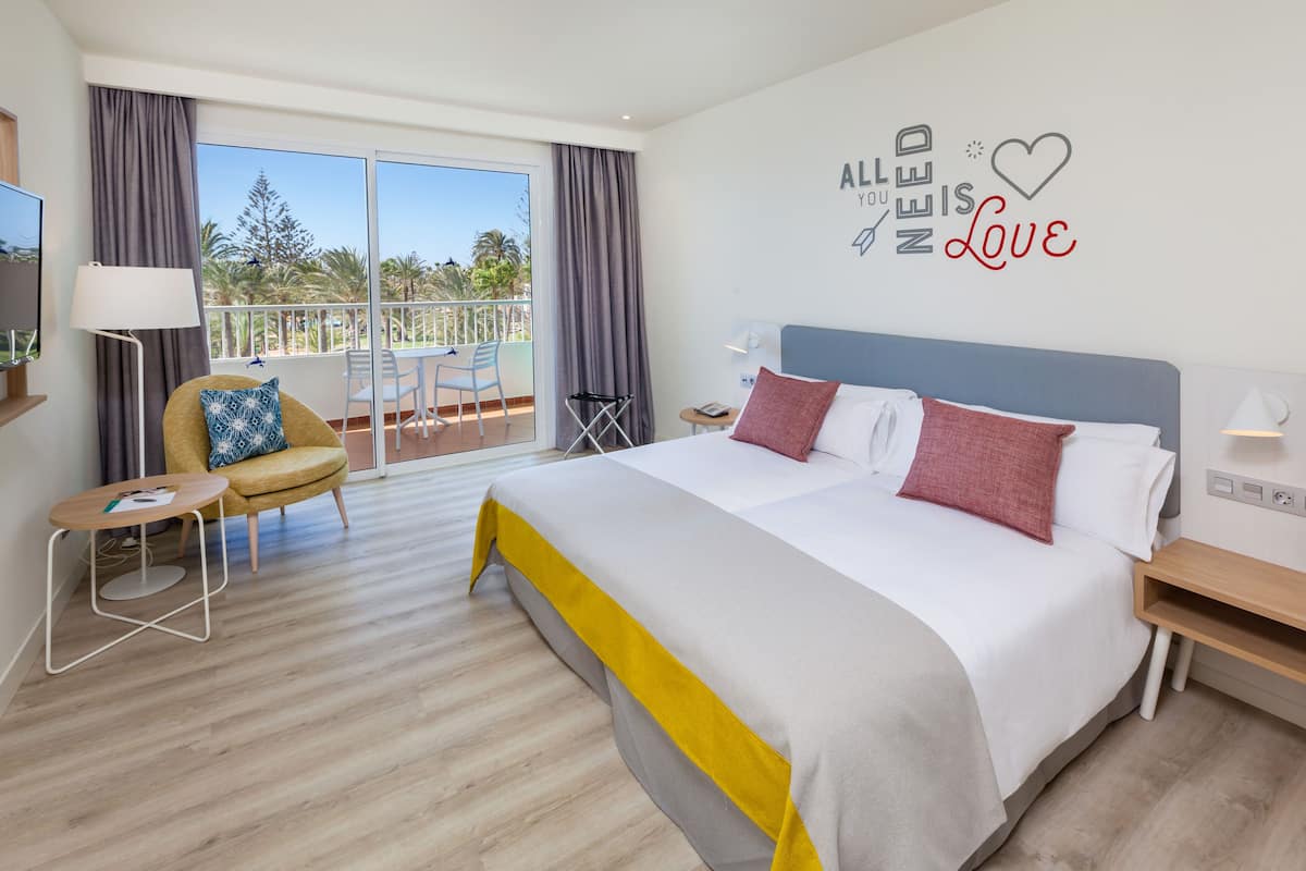 Une chambre d’hôtel moderne avec un grand lit, une petite table, une chaise et un balcon offrant une vue sur les palmiers.