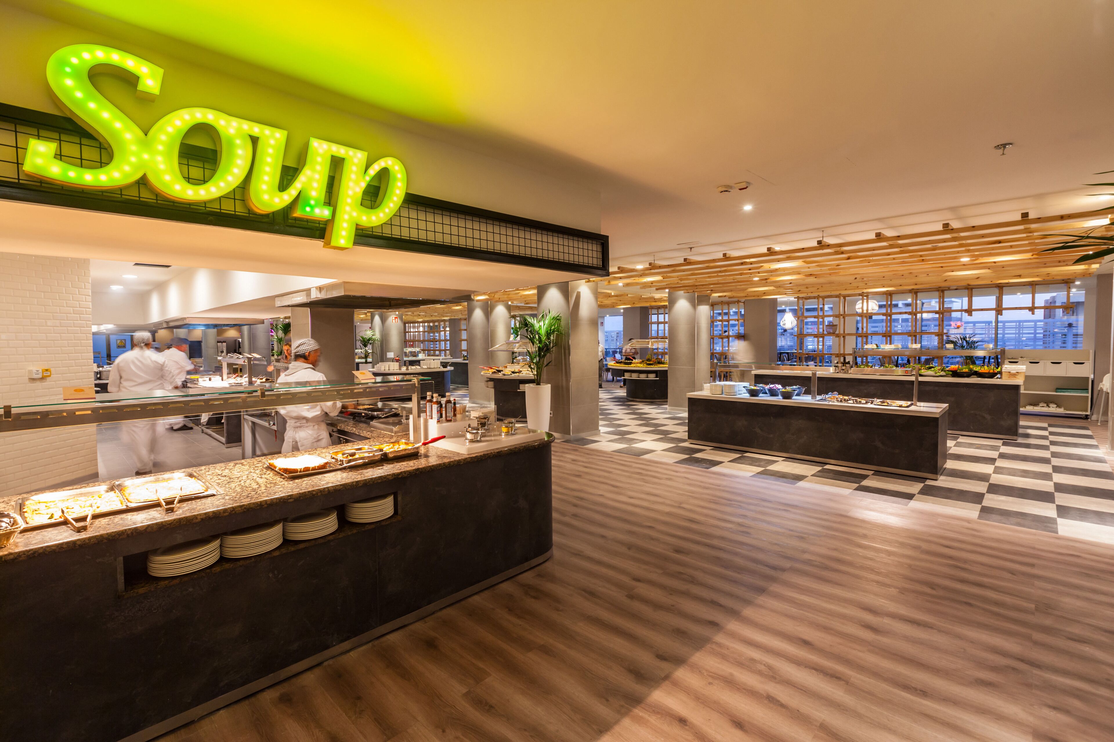 A modern restaurant interior with a prominent 'Soup' sign, a checkered floor, and a view of the city through large windows.