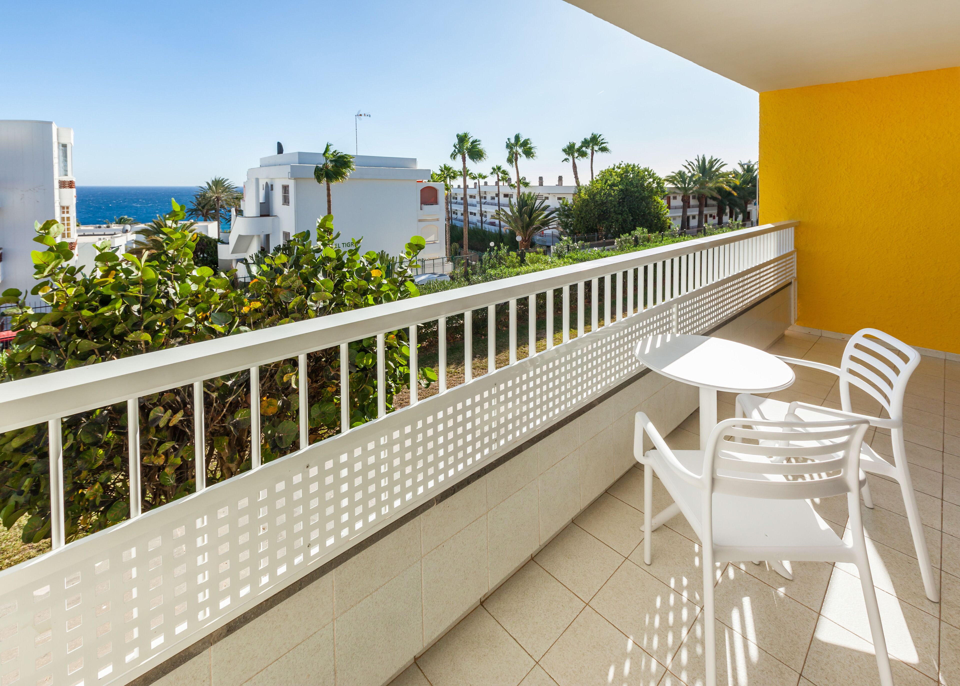 Ein Balkon mit weißen Stühlen und einem Tisch, der auf eine Küstenlandschaft mit Gebäuden und Palmen blickt.