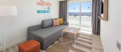 A modern hotel room with a grey sofa, orange ottomans, a small wooden coffee table, and a large window offering a view of the beach.