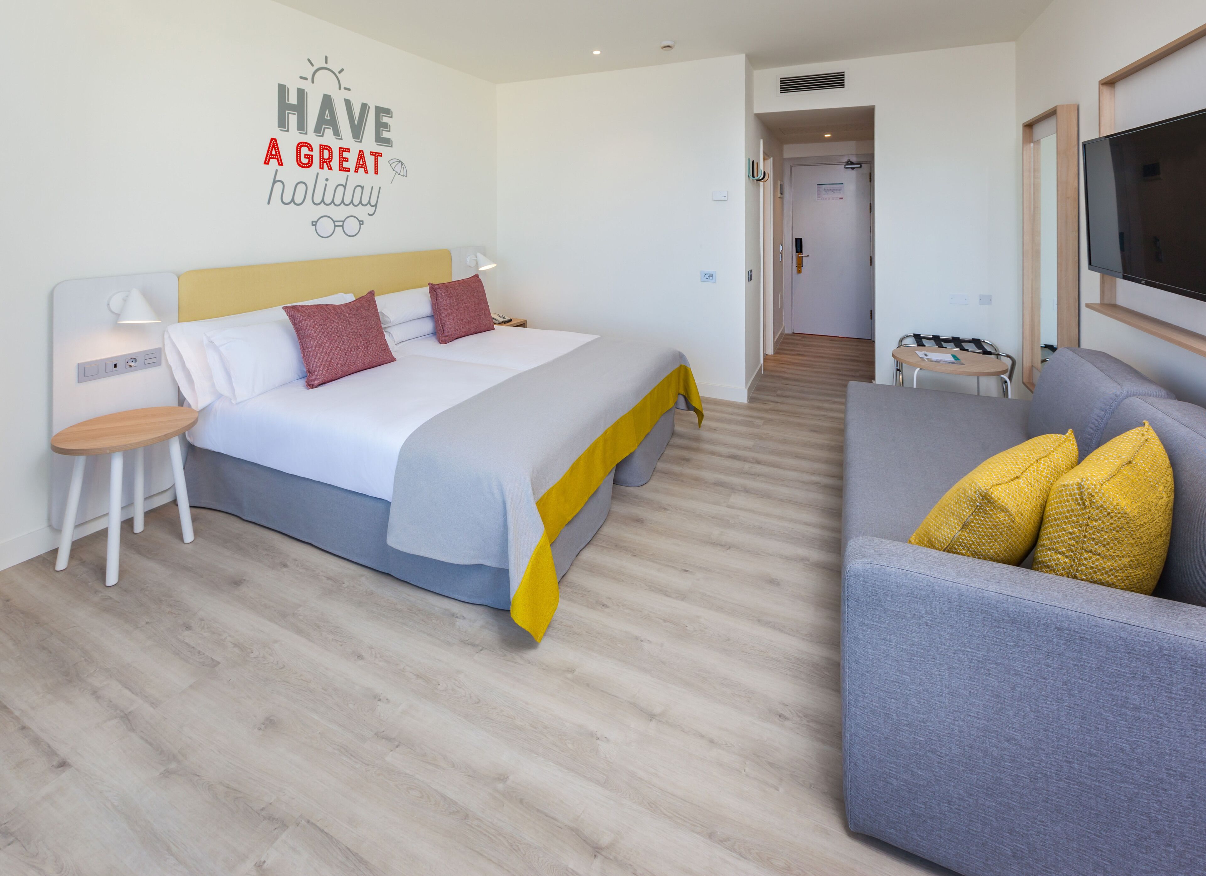 A modern hotel room with a bed, a bedside table, a chair, a sofa, and a TV.