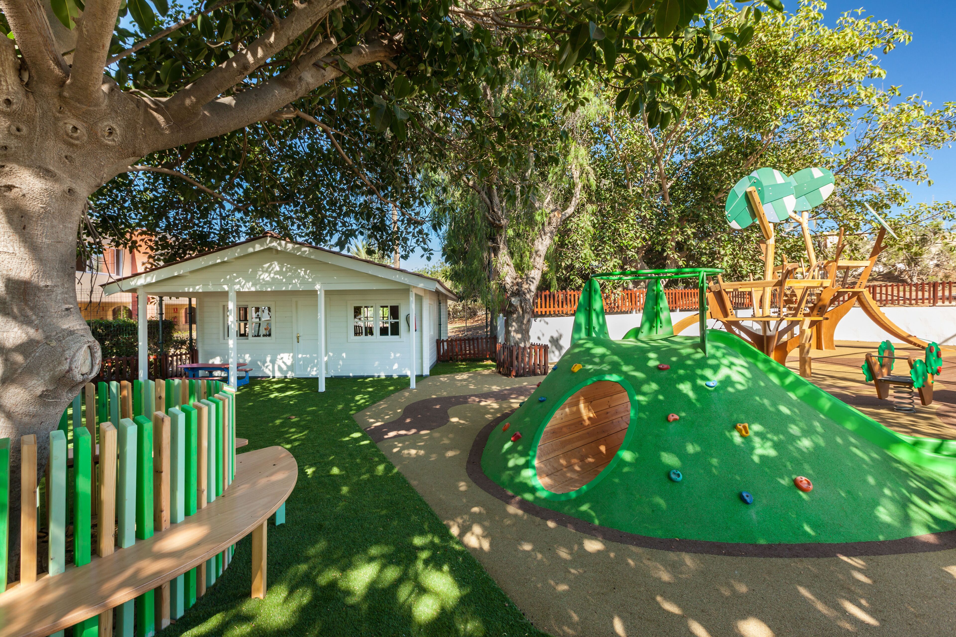 Ein Spielplatz mit Klettergerüsten und einer Rutsche, umgeben von einem eingezäunten Bereich und einem Gebäude mit weißer Fassade.
