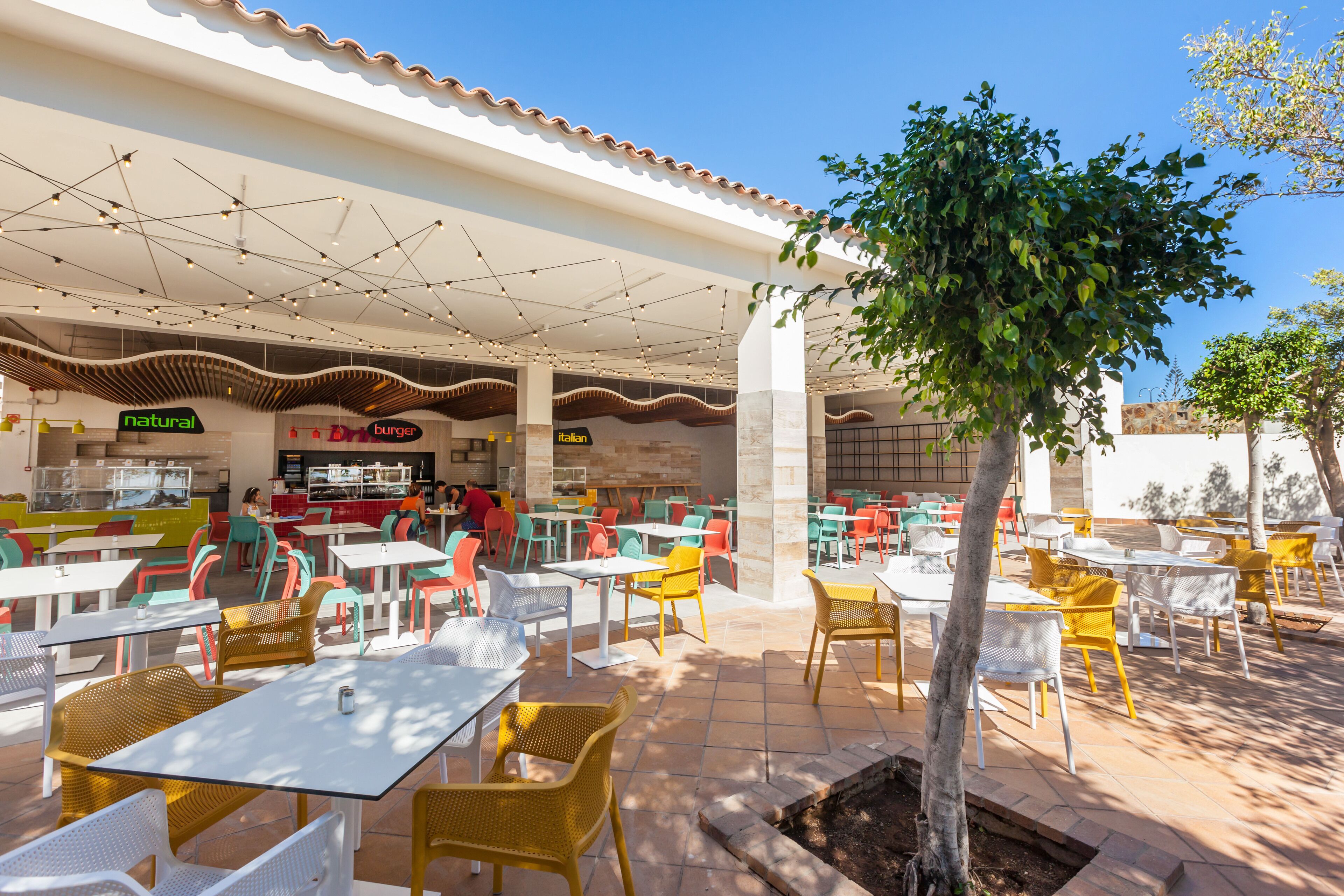 Un café en plein air avec des tables et des chaises, un arbre et un bâtiment sur lequel on peut voir des enseignes indiquant la présence de restaurants proposant une cuisine « naturelle » et « italienne ».