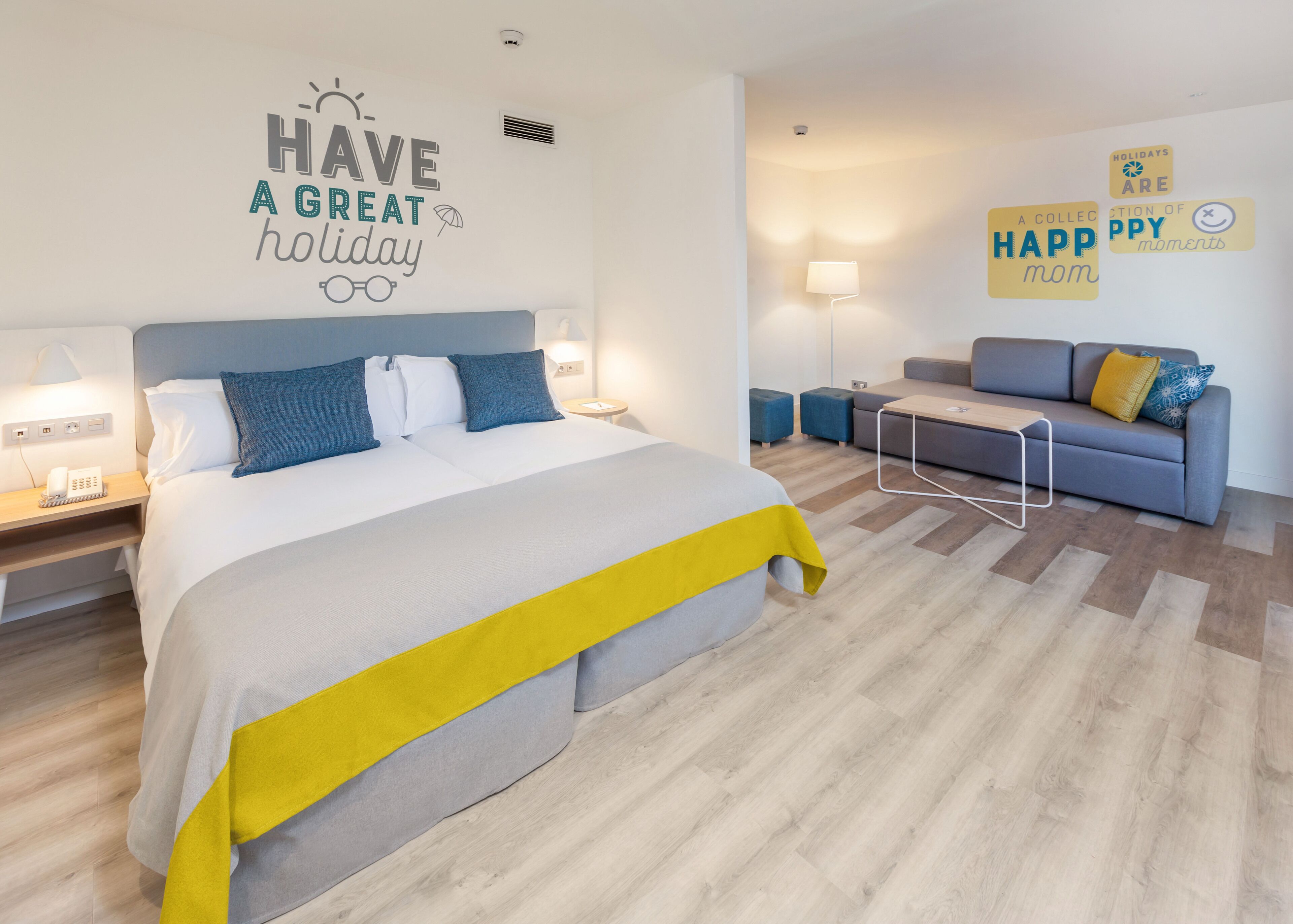A modern hotel room with a large bed, a sofa, and a desk. There are wall decorations with holiday messages.
