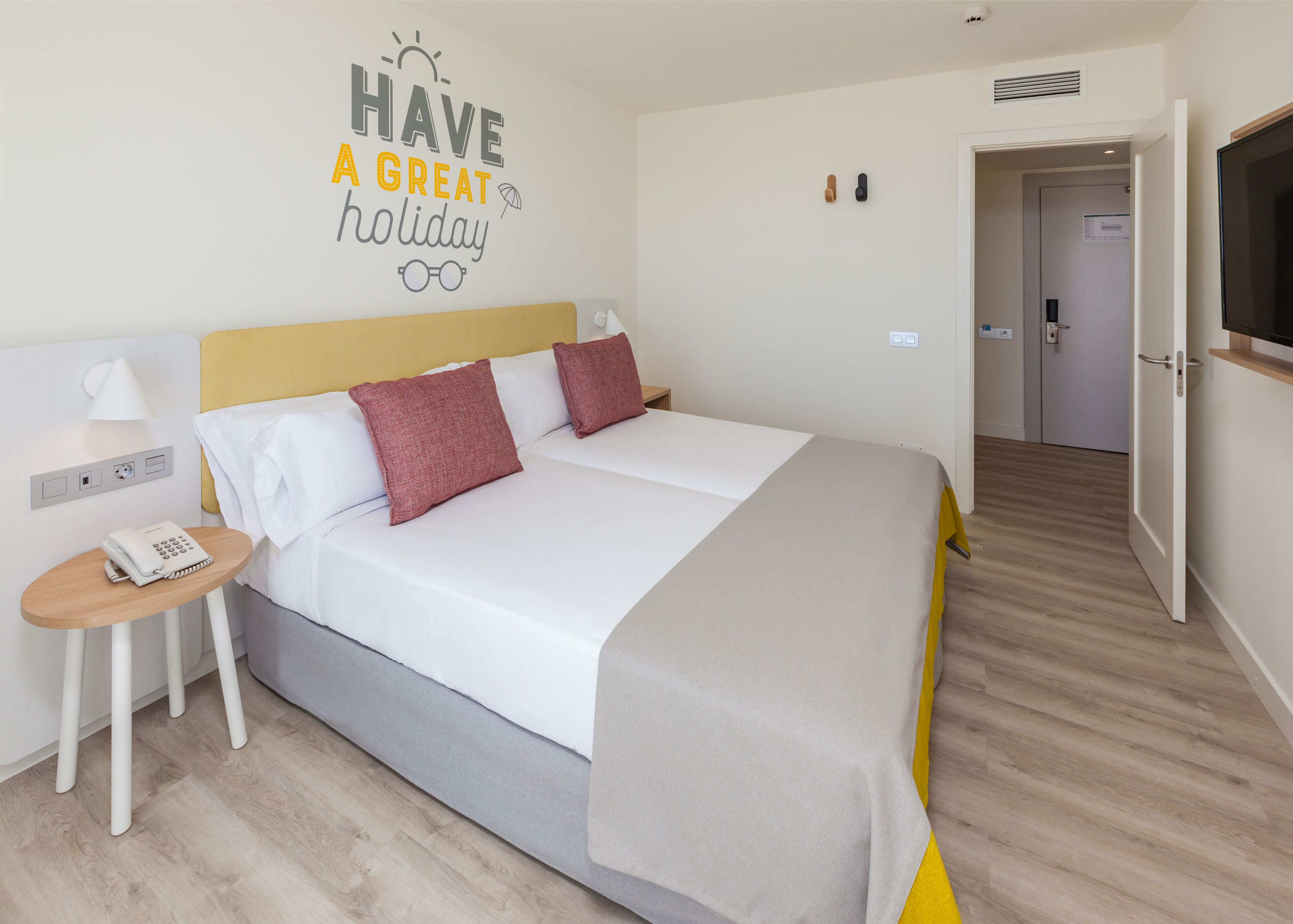 A hotel room with a bed, a bedside table with a phone, a wall-mounted lamp, and a door leading to another room.