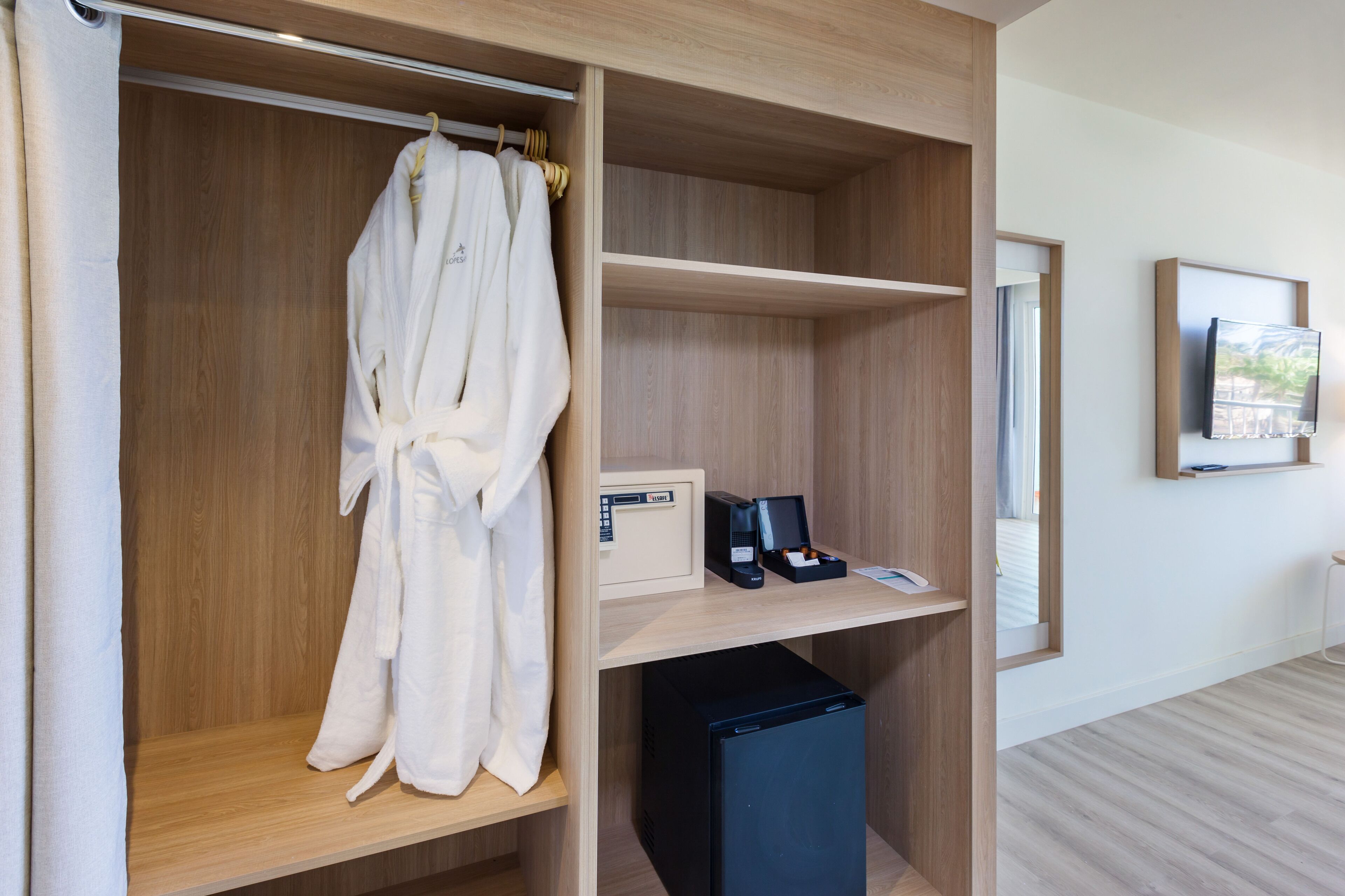 A hotel room with a wooden wardrobe, a robe hanging inside, a small desk with a phone and a coffee machine, and a TV mounted on the wall.